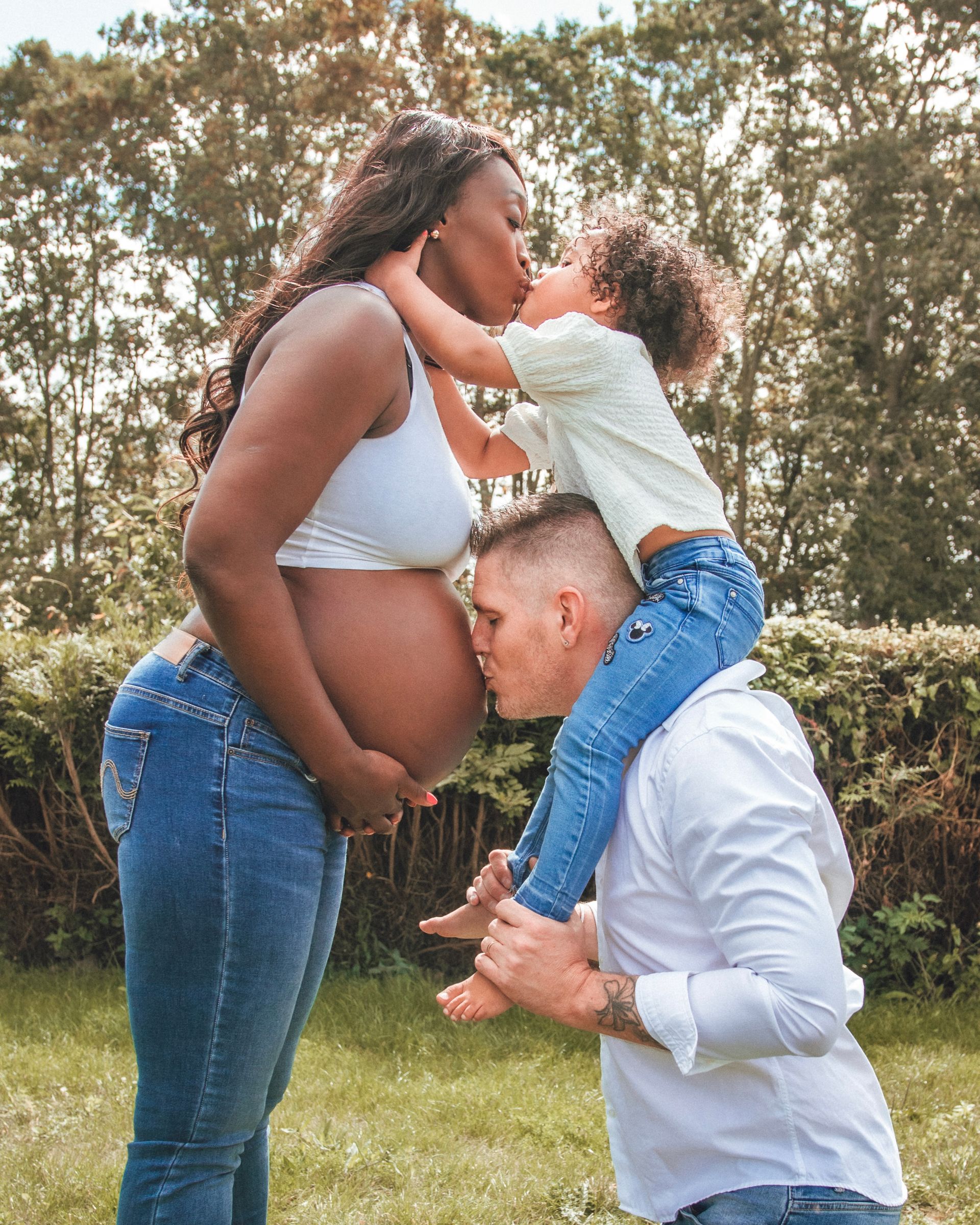 Femme noire enceinte en jean, haut blanc et un enfant l'embrassant sur la joue, tandis que le père embrasse son ventre.