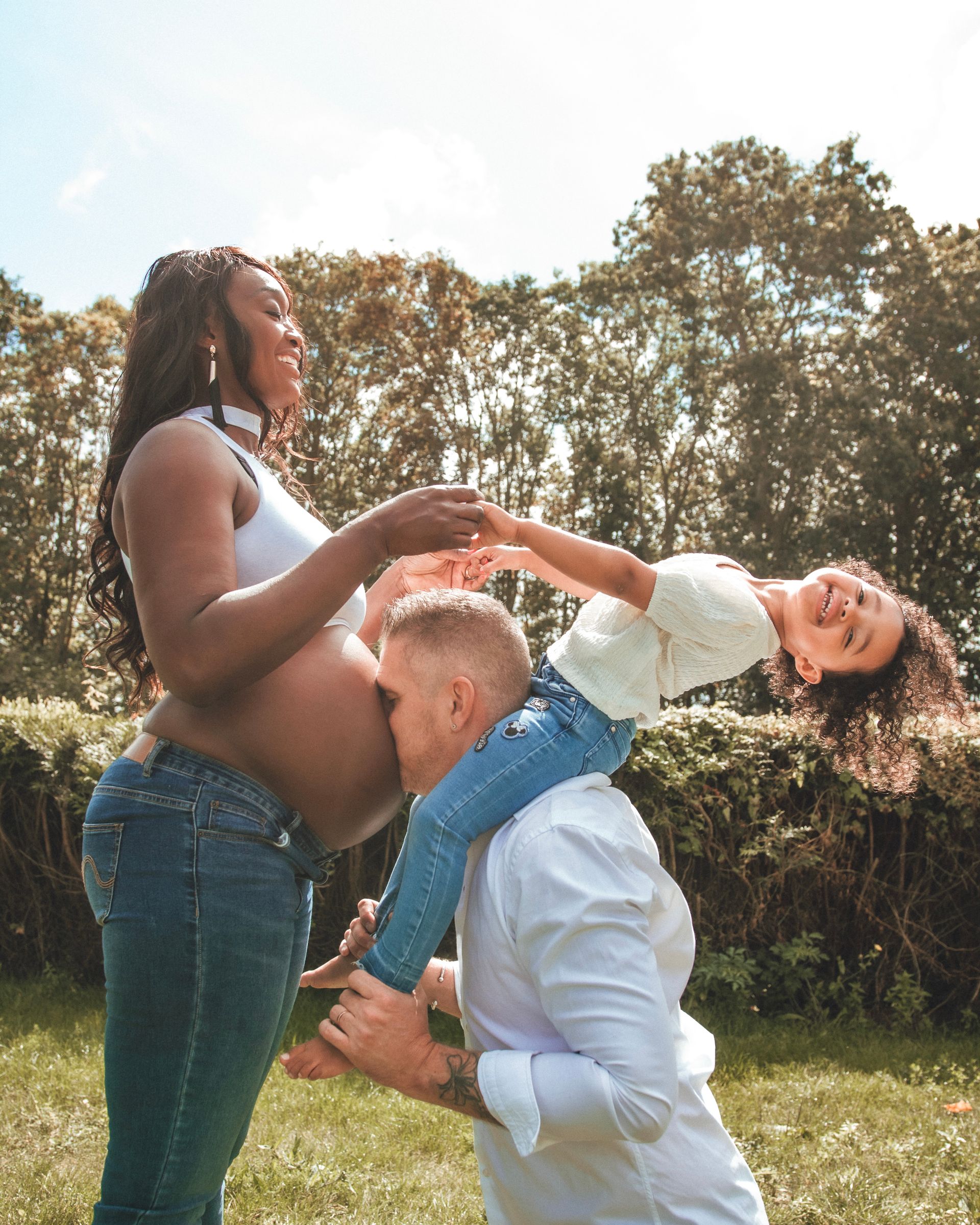 Femme enceinte avec son mari embrassant son ventre, sa fille sur ses épaules, à l'extérieur.