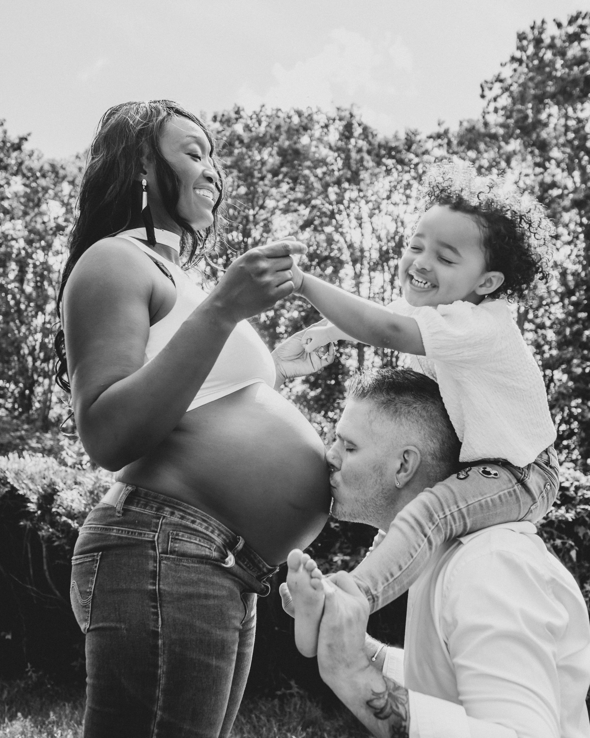 Femme enceinte avec son enfant sur les épaules de son père. Le père embrasse son ventre. Expressions joyeuses en extérieur.