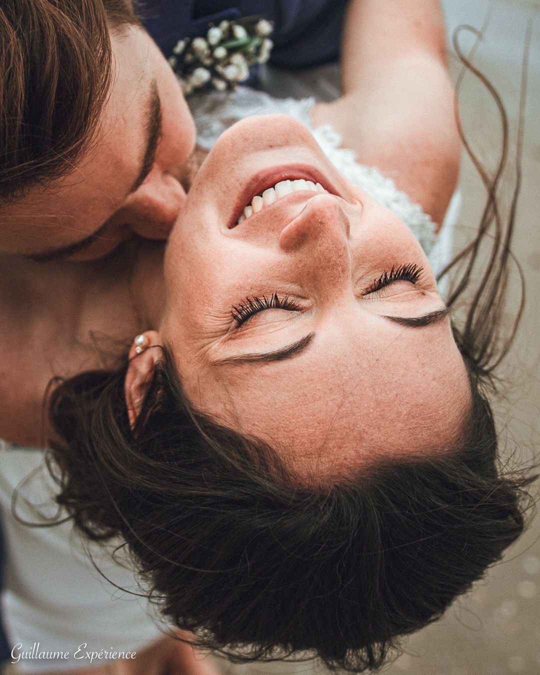 Un homme embrasse le cou d'une femme ; elle sourit, les yeux fermés. Lumière douce, léger flou.