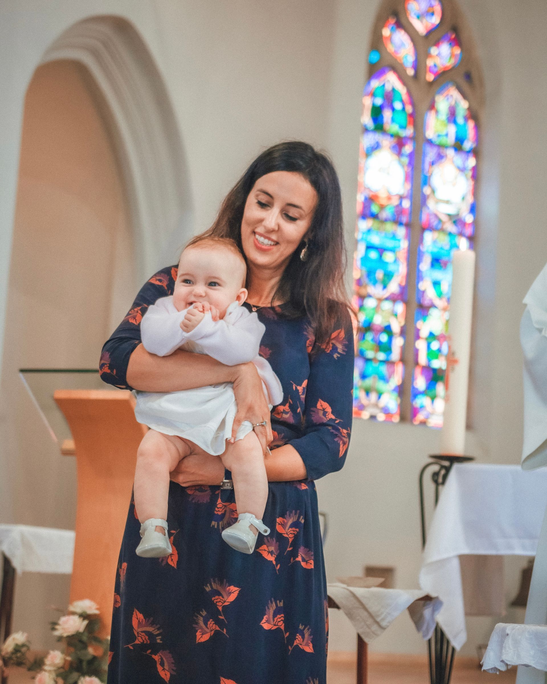 Une femme en robe bleue tient un bébé en tenue blanche dans une église avec un vitrail.
