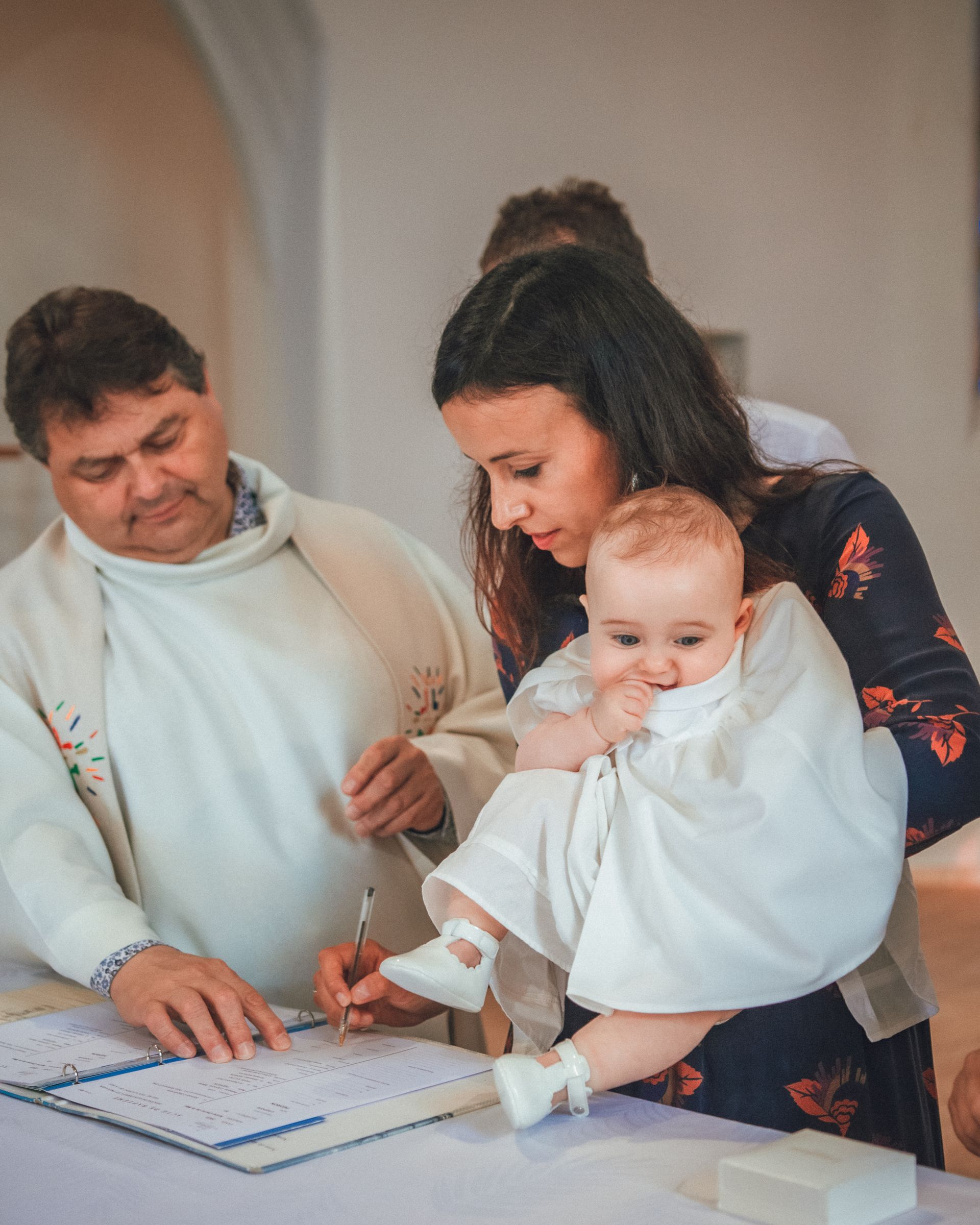 Un prêtre signe un livre tandis qu'une femme tient un bébé en robe blanche lors d'un baptême.