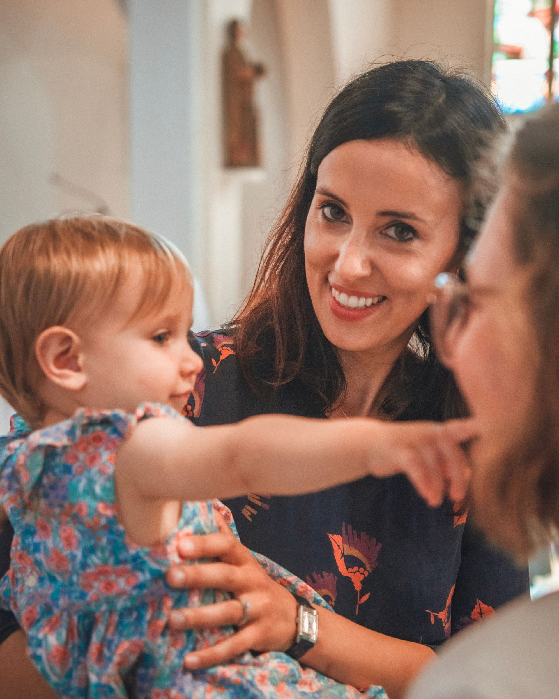 Une femme sourit, tenant un enfant en bas âge, et désigne une autre personne dans une église.