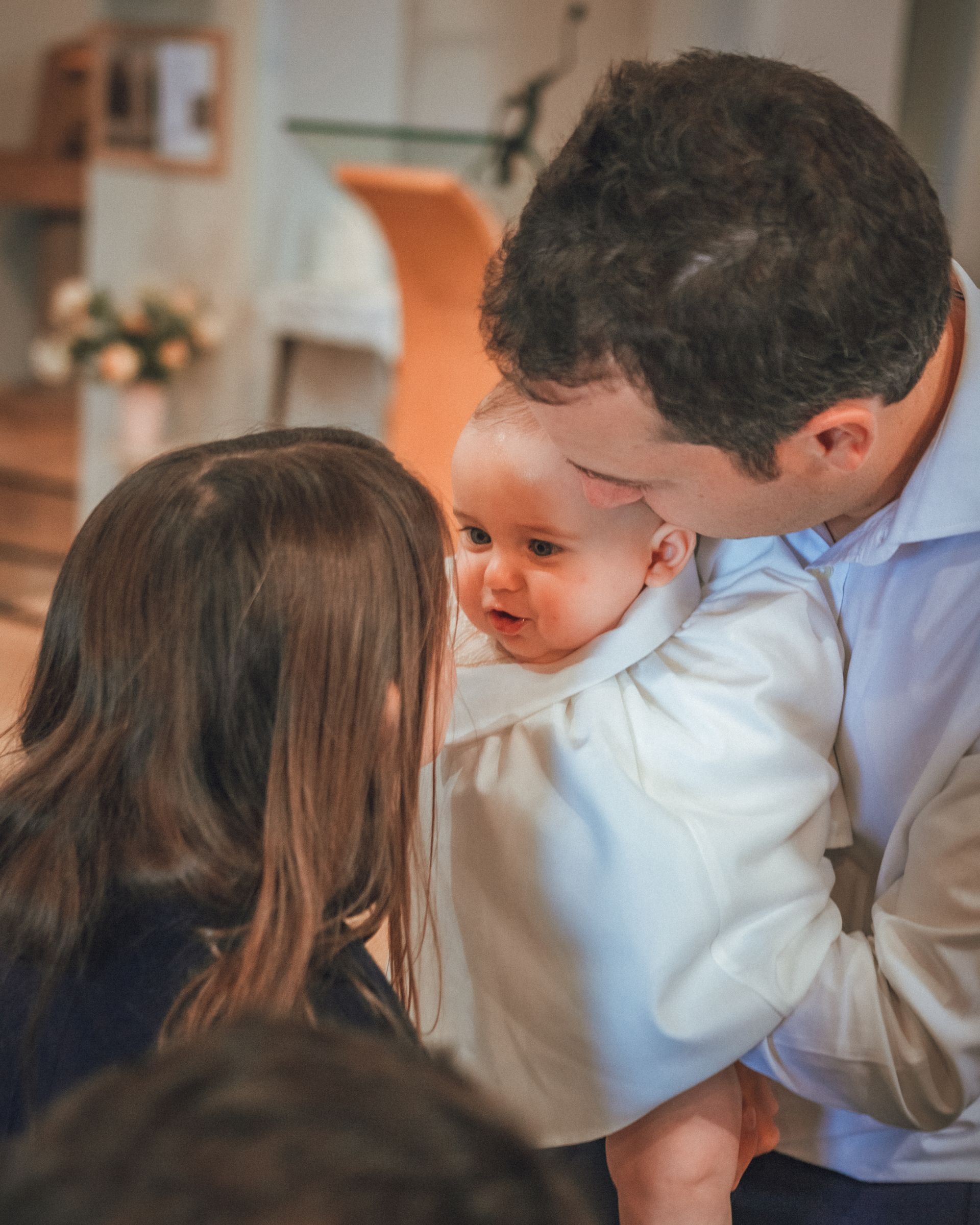 Homme tenant un bébé dans une robe blanche, regardant une femme ; intérieur de l'église.