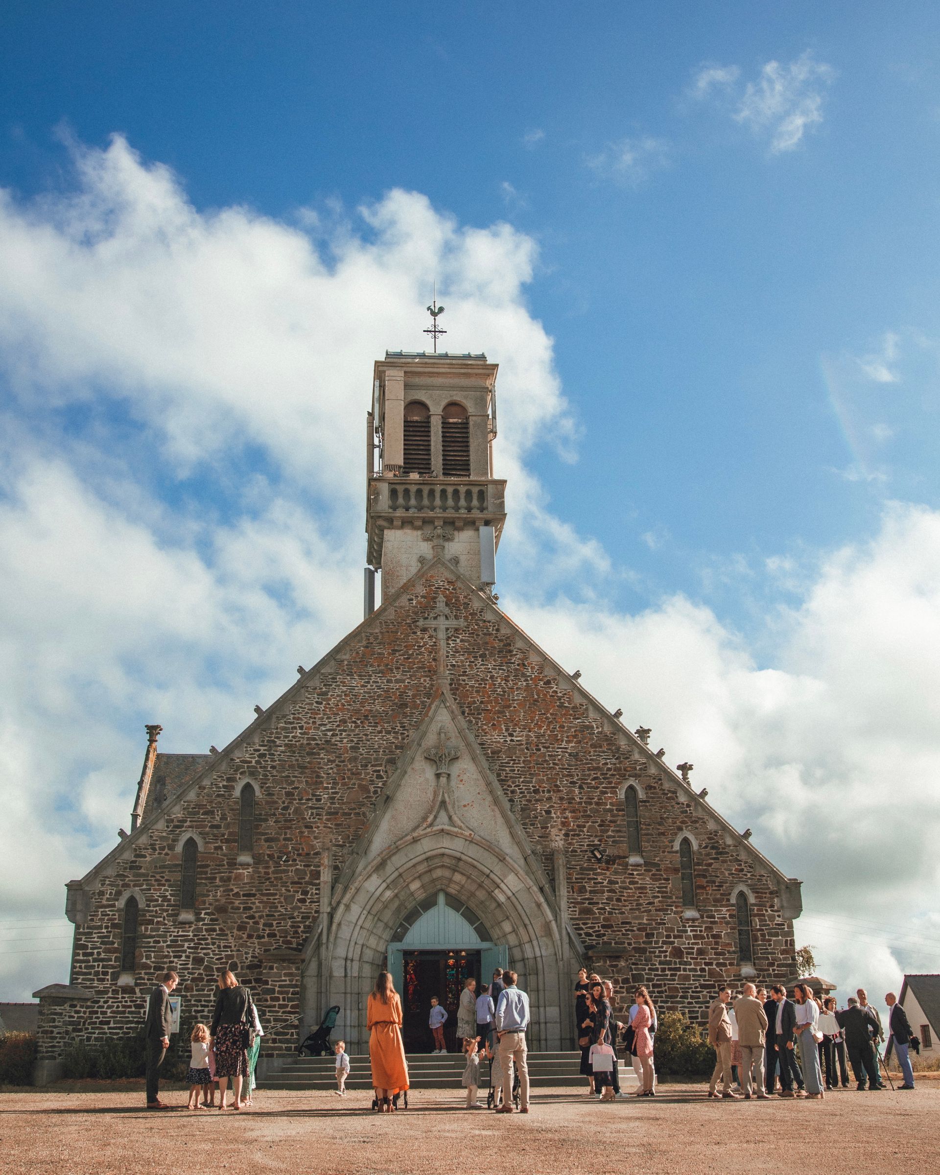 Extérieur de l'église avec des gens rassemblés, journée ensoleillée. Clocher avec cloches.