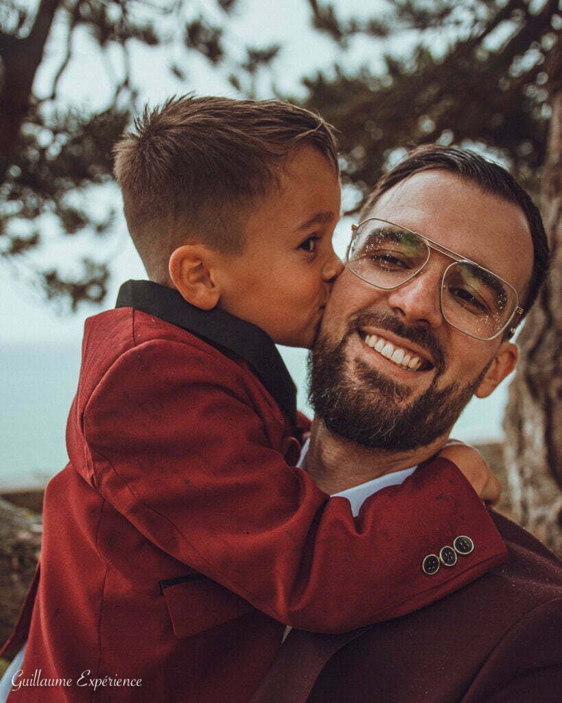 Un jeune garçon en costume rouge embrasse son père sur la joue. Ils sont dehors, souriants.