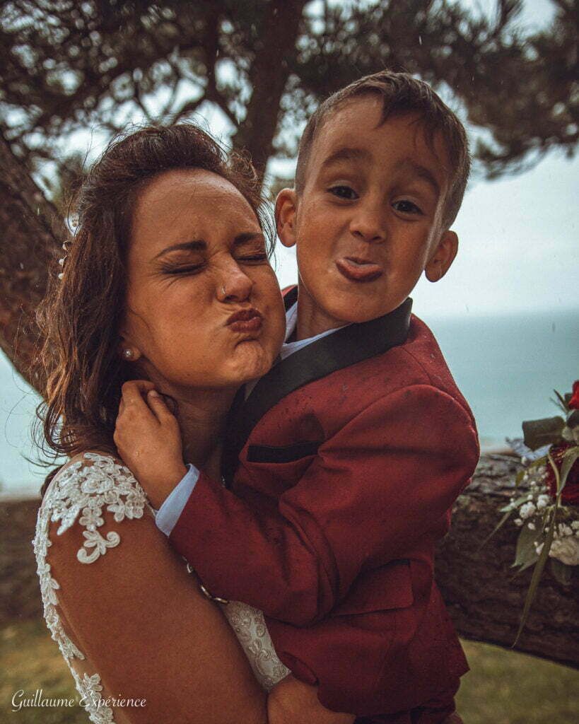 La mariée et l'enfant font des grimaces à l'extérieur ; la mariée en robe de dentelle, l'enfant en costume rouge.