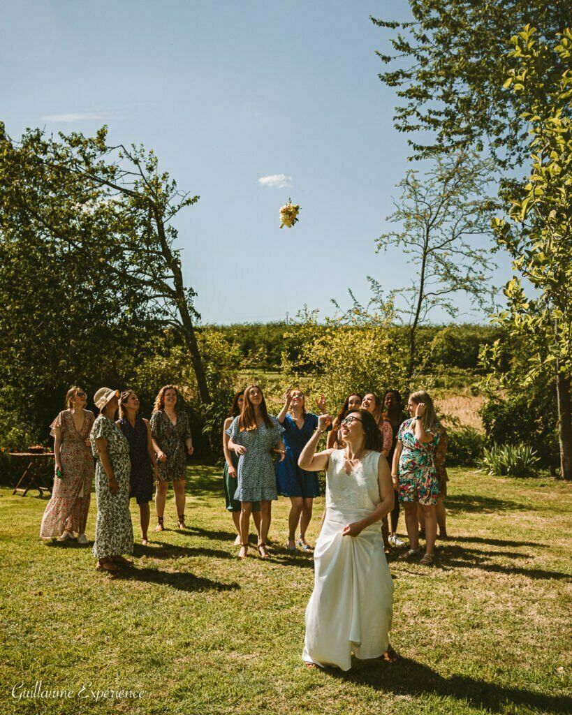 Une mariée lance un bouquet à un groupe de femmes dans un cadre extérieur ensoleillé avec des arbres.