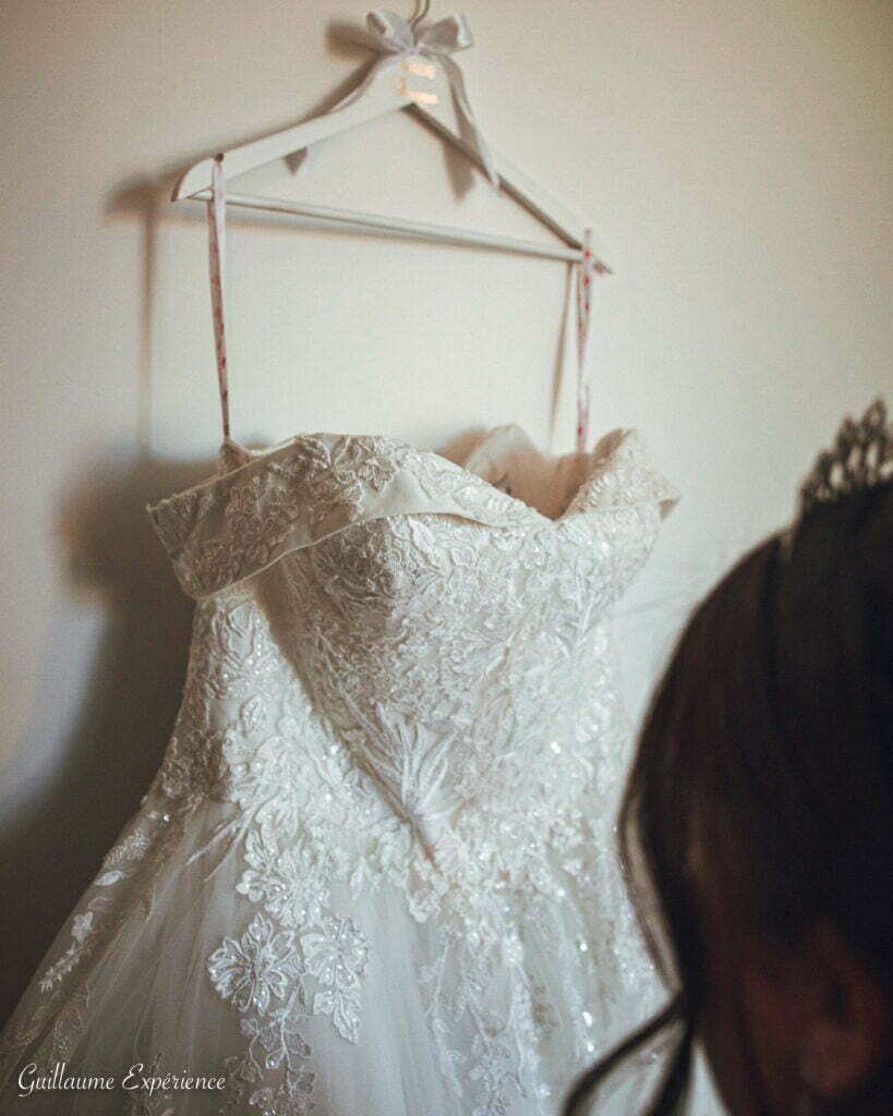 Robe de mariée suspendue à un cintre en bois ; une mariée regarde, un diadème visible.