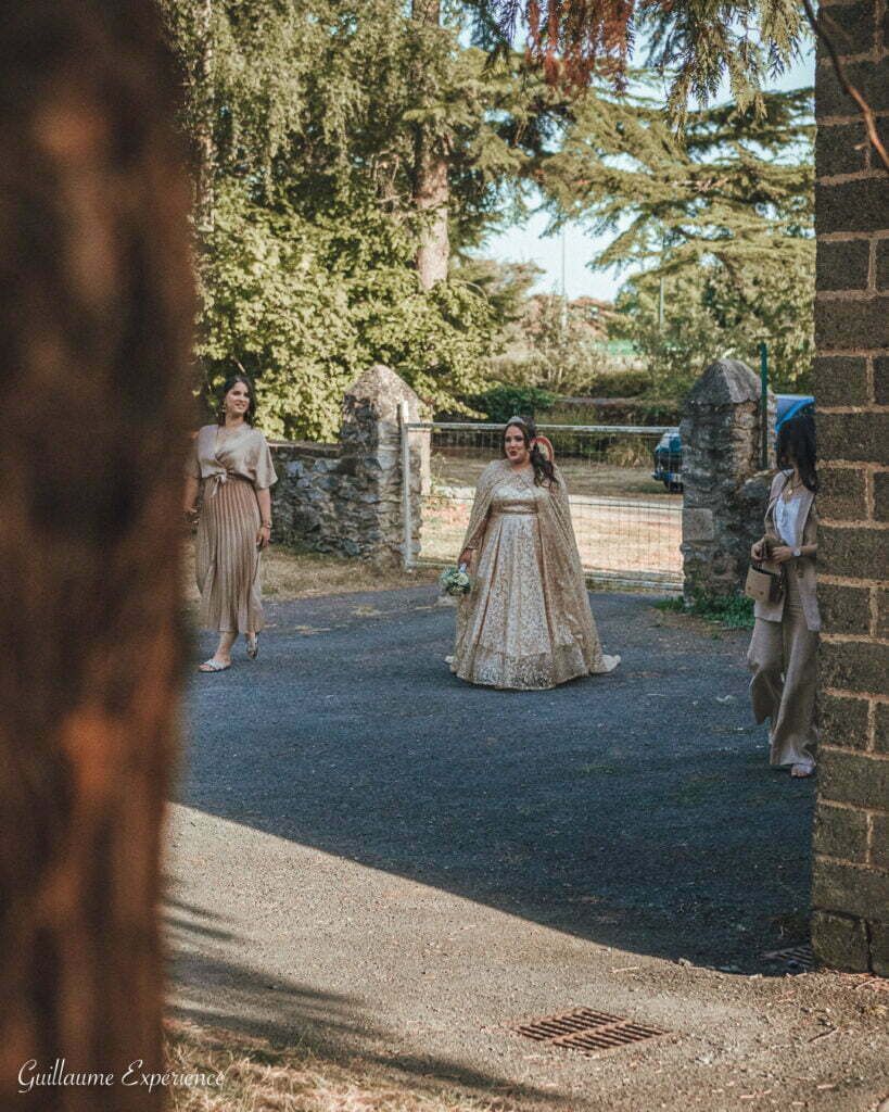 La mariée en robe dorée marche vers la caméra avec deux demoiselles d'honneur près d'un mur de pierre et d'un portail.