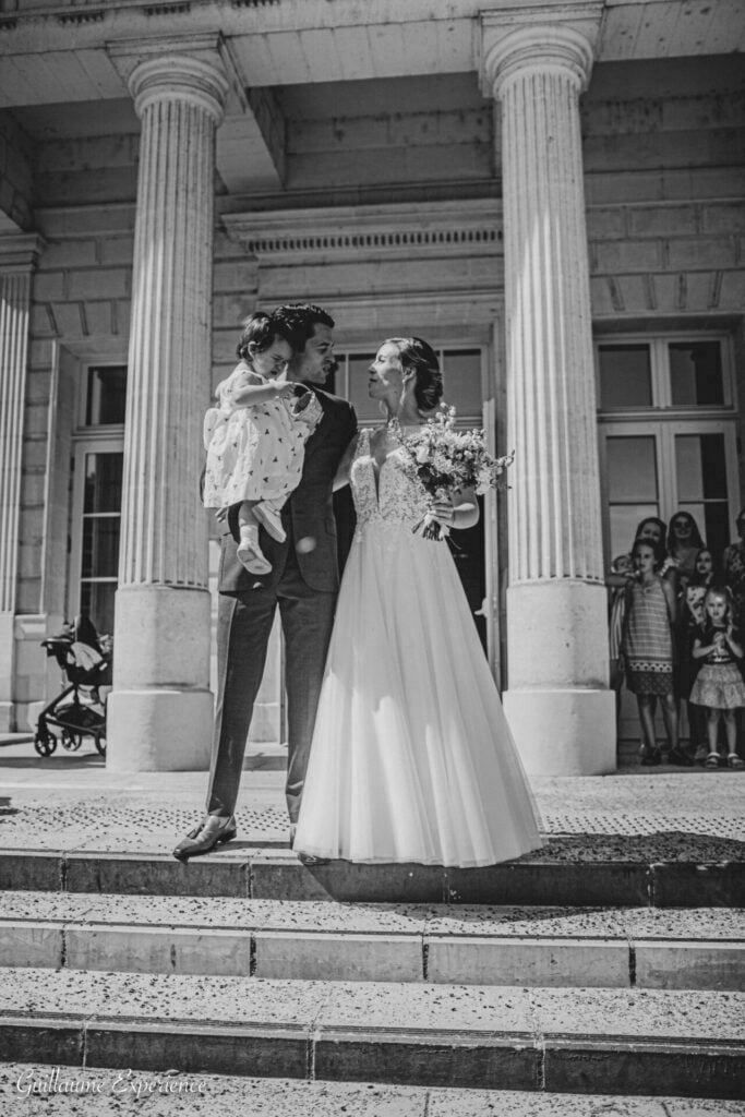 Des jeunes mariés se tiennent devant un bâtiment avec un enfant, tenant un bouquet, en noir et blanc.