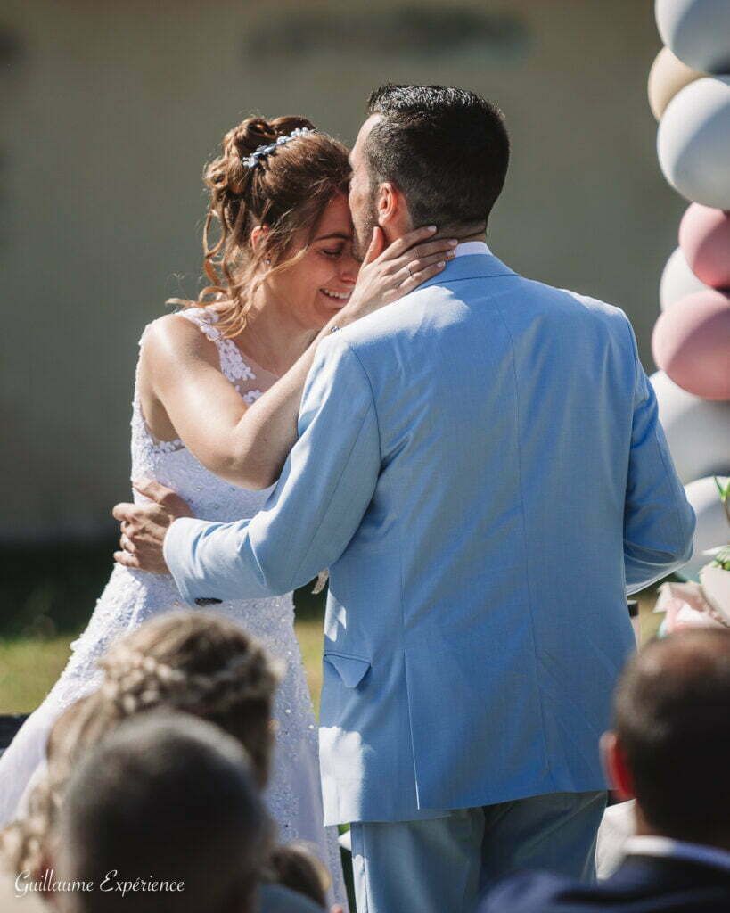 Les mariés s'embrassent lors de leur cérémonie de mariage ; costume bleu clair, robe blanche, décor extérieur.