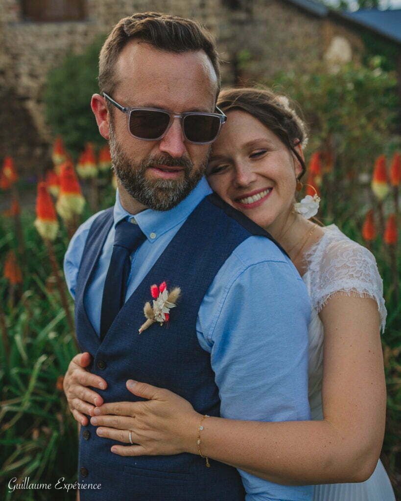 Les jeunes mariés s'embrassent en plein air : l'homme en gilet, lunettes de soleil, la femme en robe blanche sourit.