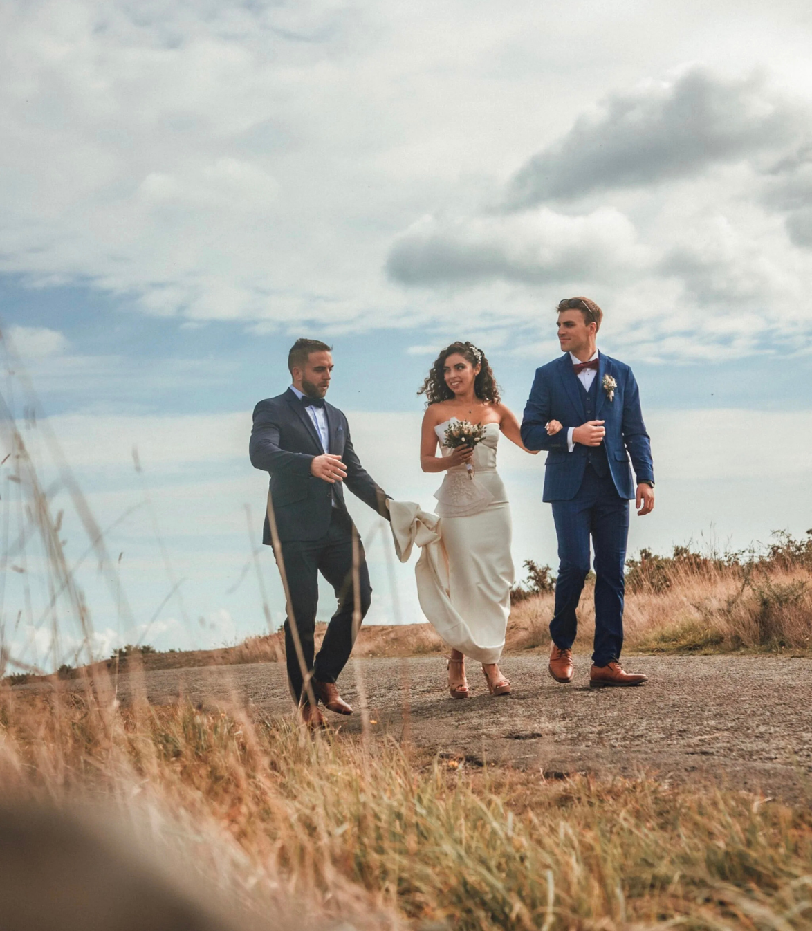 La mariée marche entre deux garçons d'honneur sur un chemin de terre. Cadre extérieur ensoleillé.