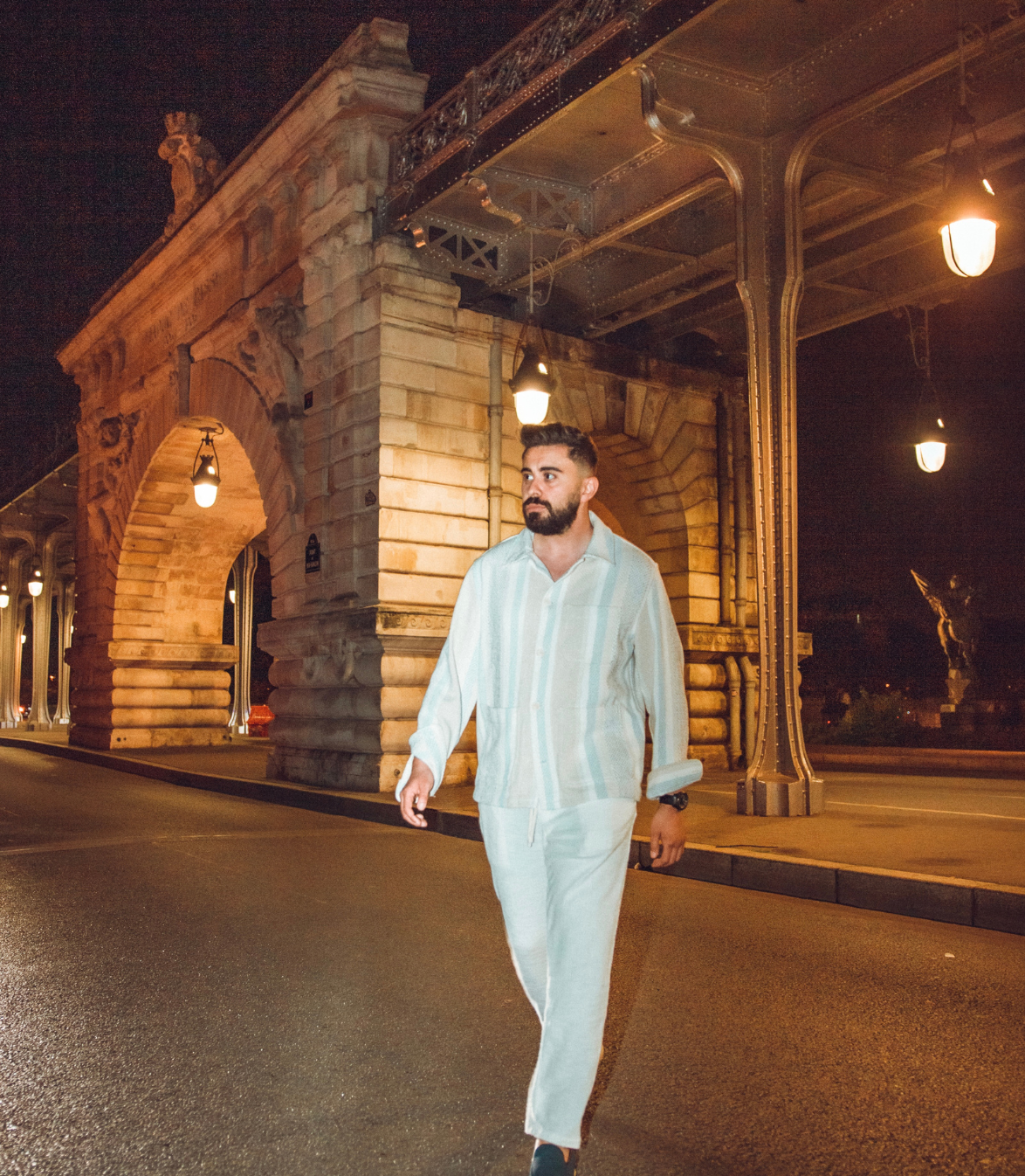 Un homme marchant sous un pont de pierre éclairé la nuit, vêtu d'une tenue en lin de couleur claire.