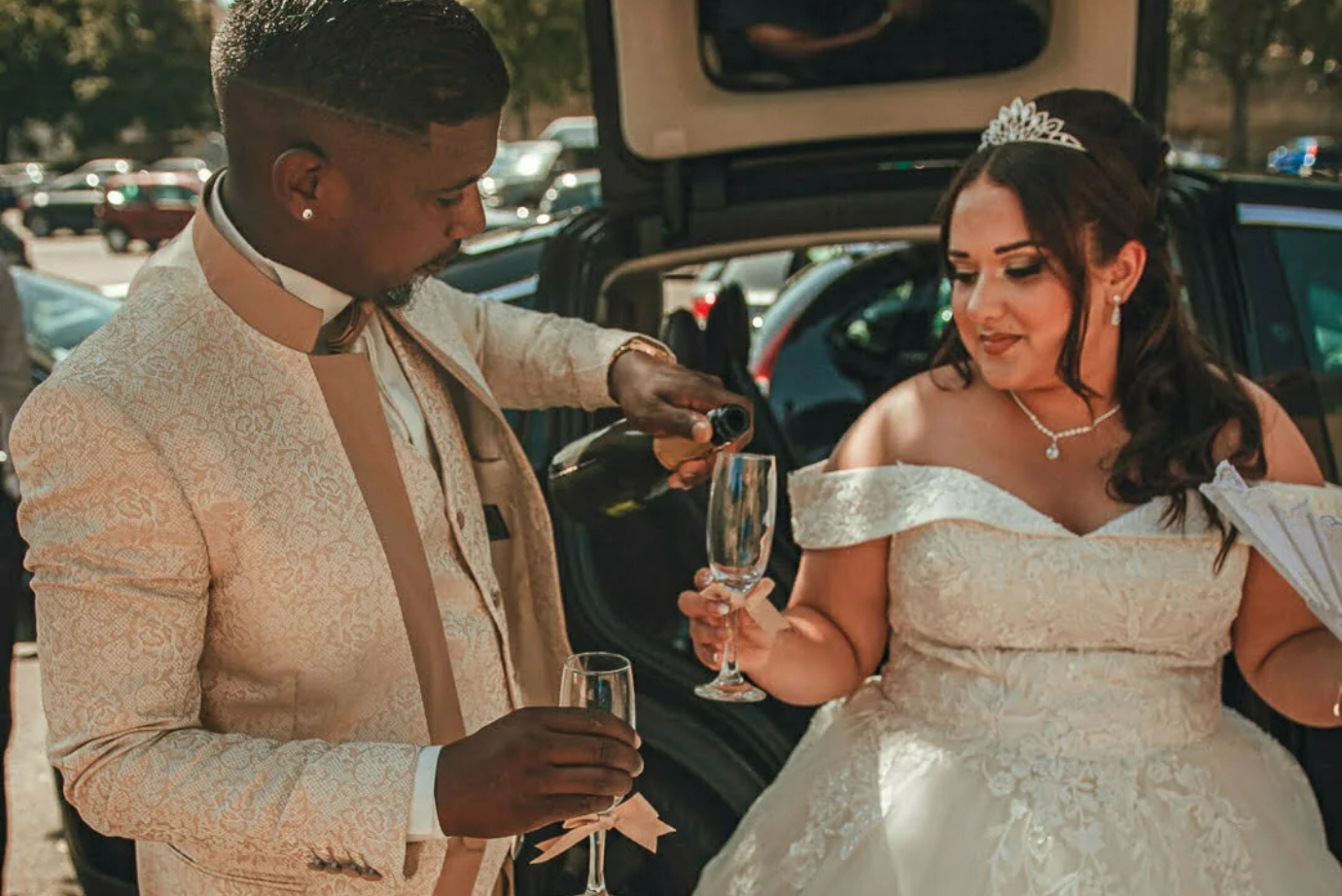 Le marié en costume crème verse du champagne à la mariée en robe à épaules dénudées en voiture.
