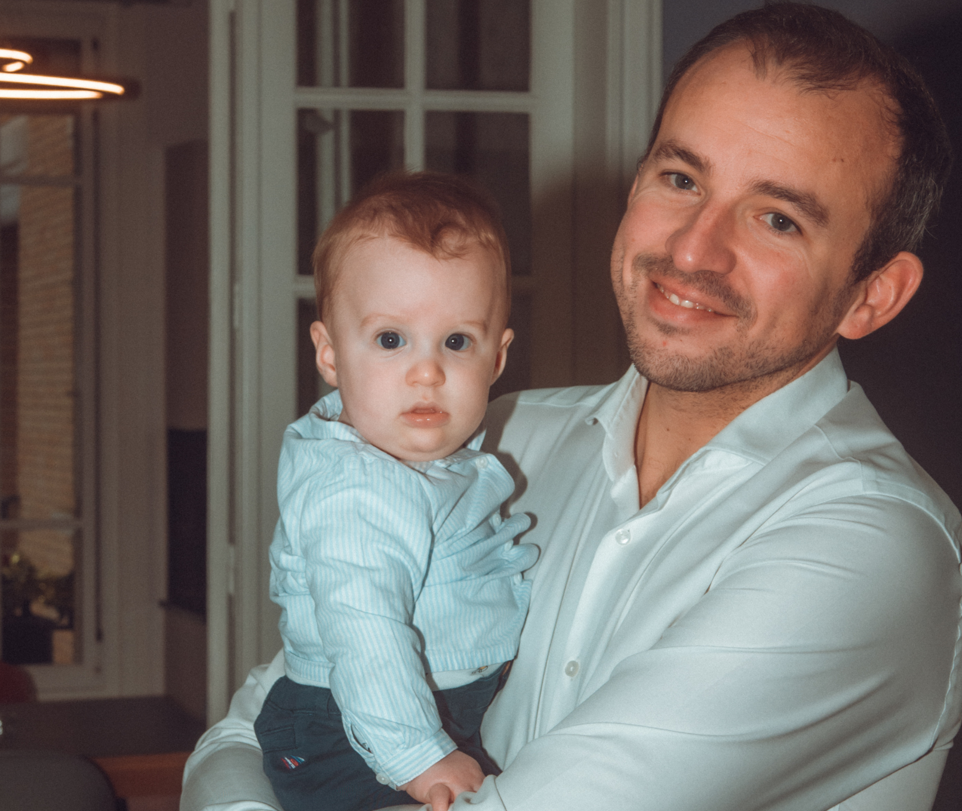Un homme tient un bébé. Tous deux sourient. Le bébé porte un t-shirt bleu clair. Dans une pièce.