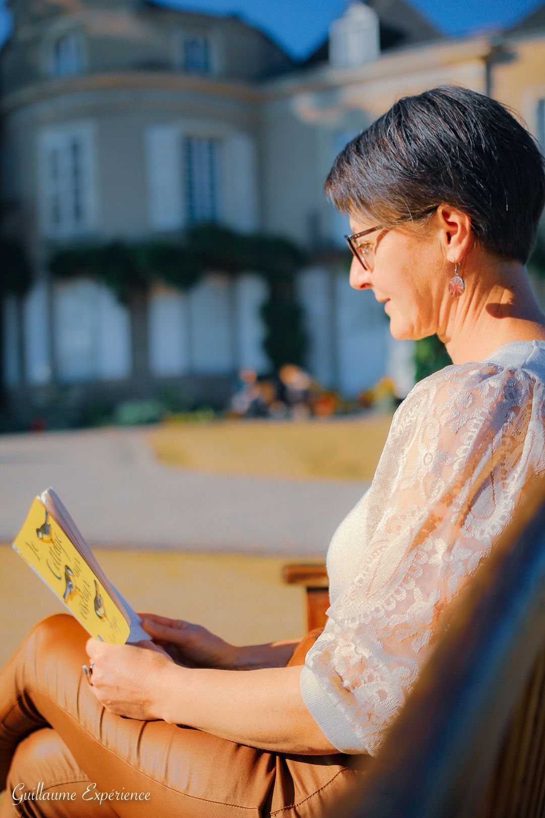 Une femme aux cheveux gris courts lit un livre à l'extérieur, pantalon marron, haut en dentelle beige, journée ensoleillée.