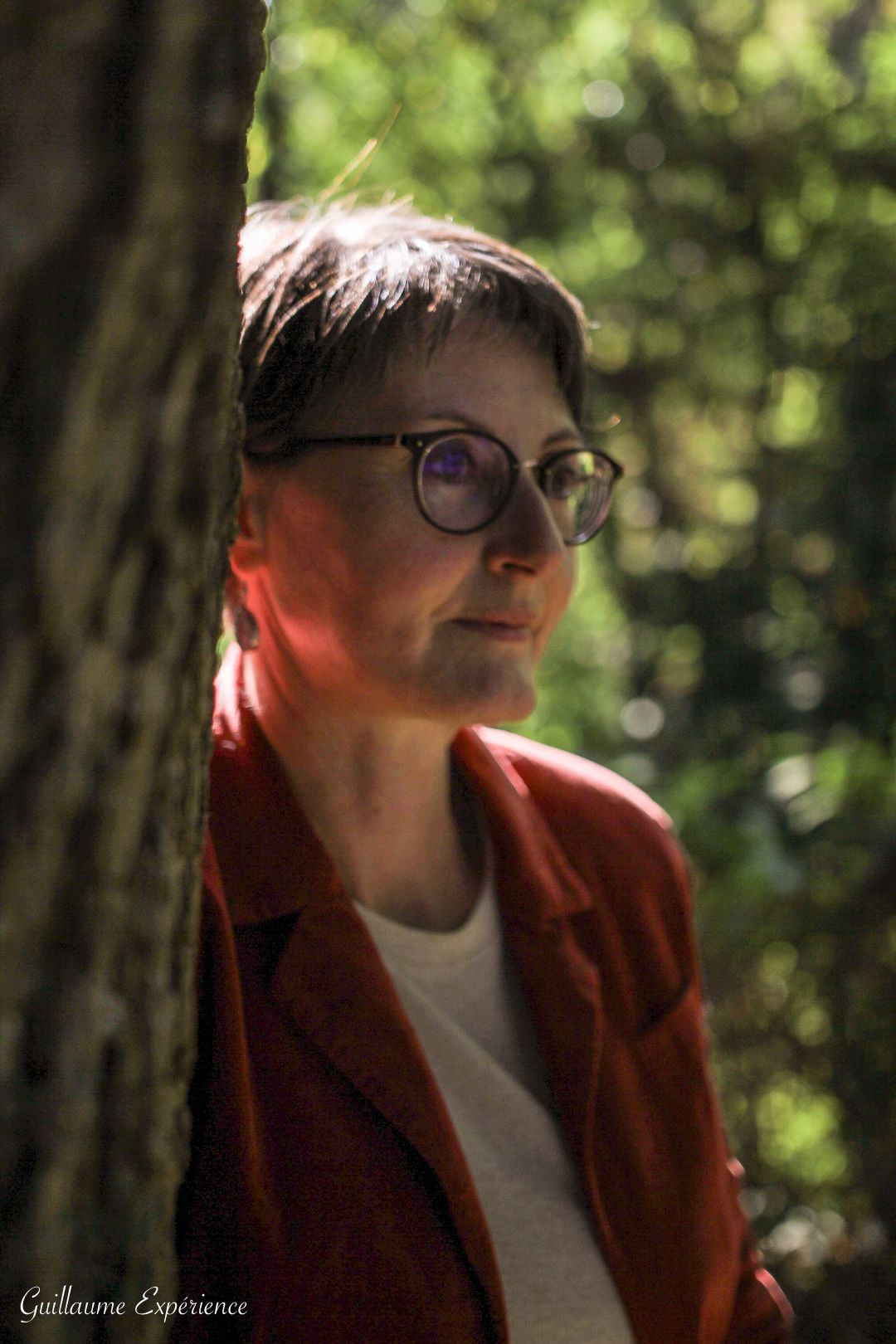 Une femme avec des lunettes et une veste rouge s'appuie contre un arbre, regardant au loin, à l'extérieur.
