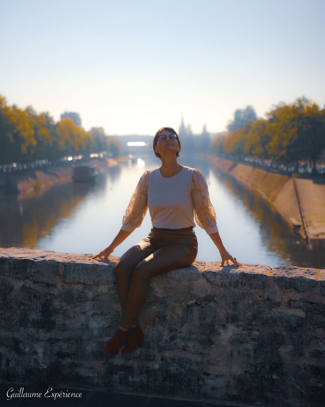 Une femme est assise sur un pont de pierre, les bras tendus, regardant le ciel, la rivière en arrière-plan.