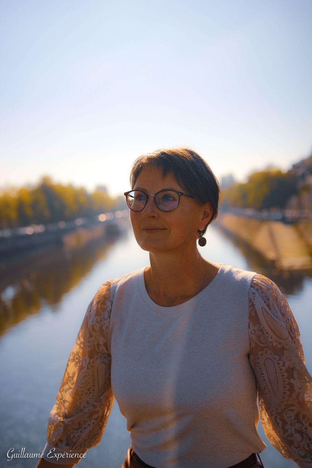 Femme avec des lunettes, cheveux courts, sur un pont, regardant sur le côté avec une rivière et des arbres en arrière-plan.
