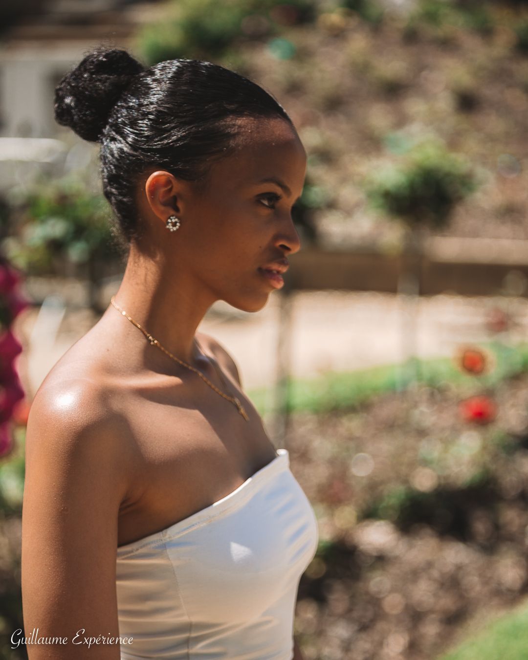 Une femme aux cheveux noirs en chignon, portant un haut tube blanc, regarde pensivement sur le côté, à l'extérieur.