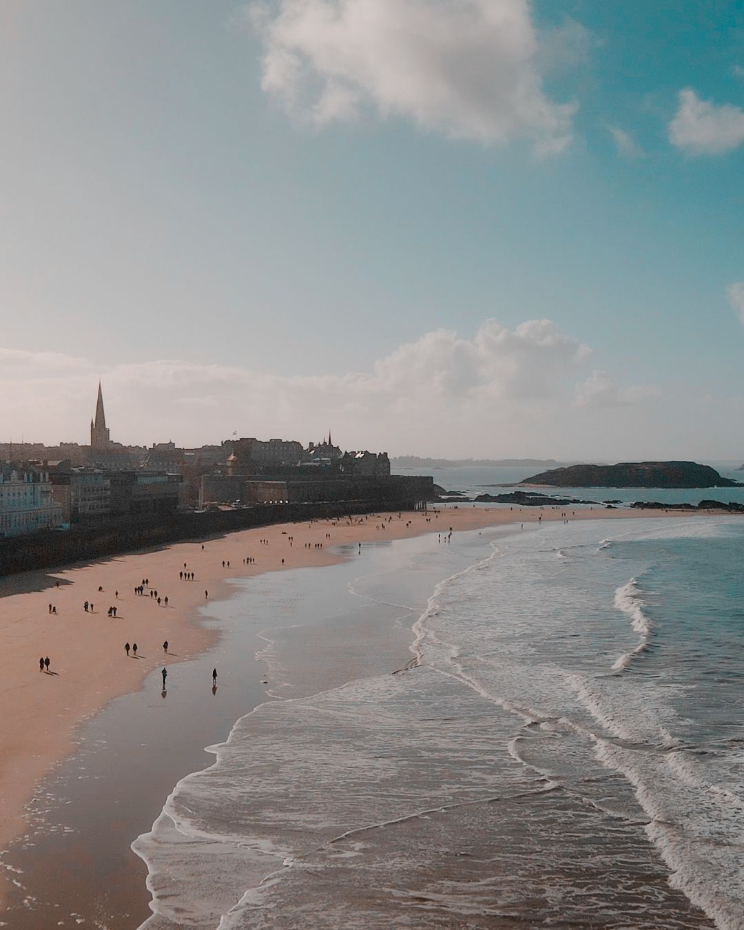 Plage de sable doré, vagues de l'océan et horizon urbain sous un ciel bleu. Des gens sont sur la plage.