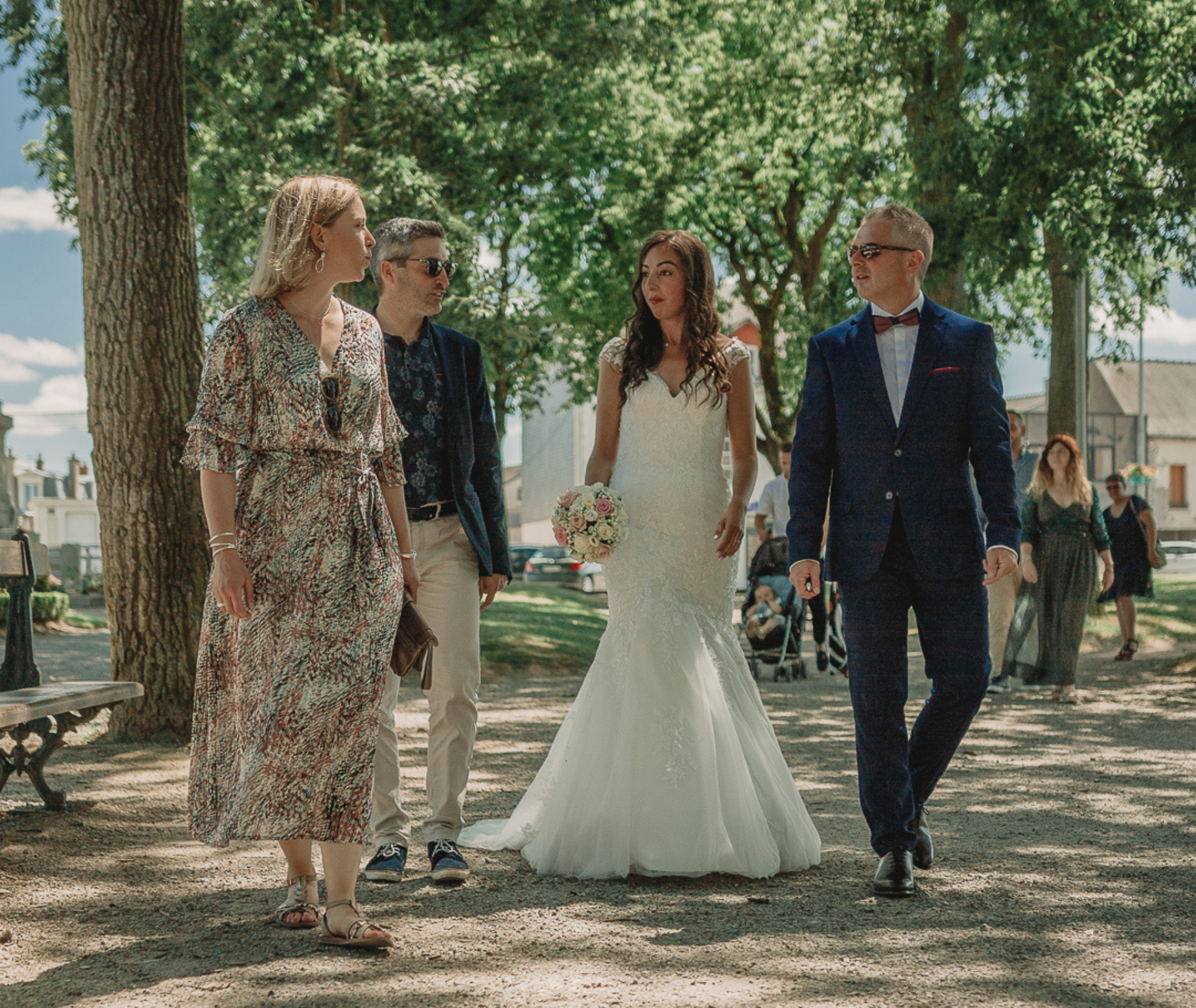 La mariée en robe blanche marche avec d'autres à l'extérieur vers un bâtiment, journée ensoleillée.