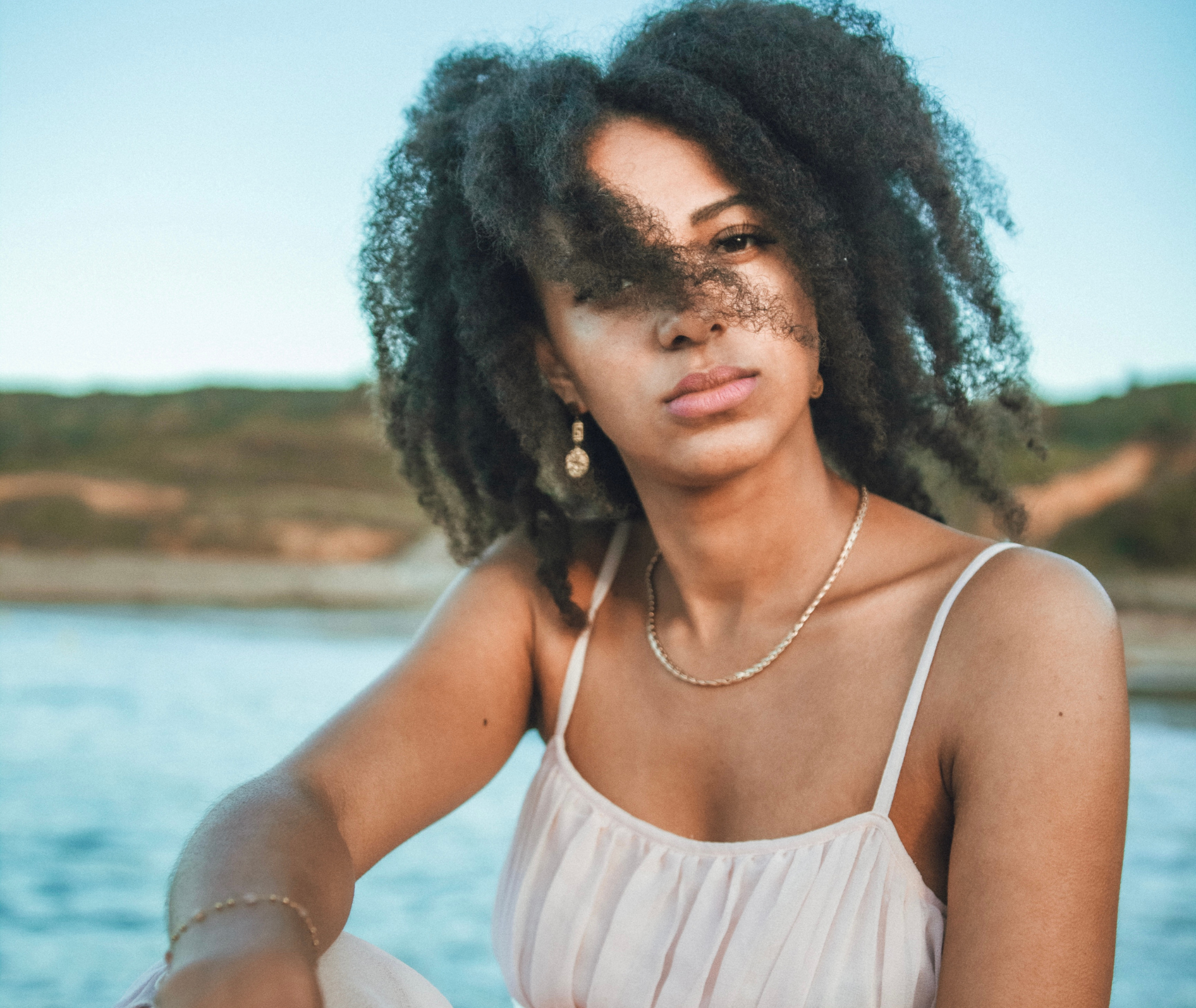 Femme aux cheveux bouclés, vêtue d'une robe blanche, près de l'eau, regardant la caméra.