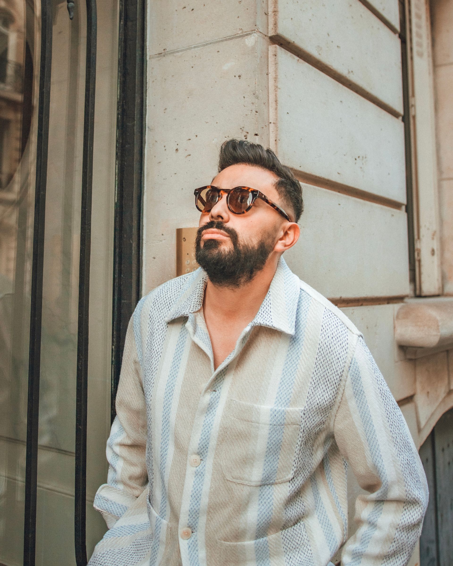 Homme avec une barbe portant des lunettes de soleil et une chemise rayée, appuyé contre un bâtiment en pierre.