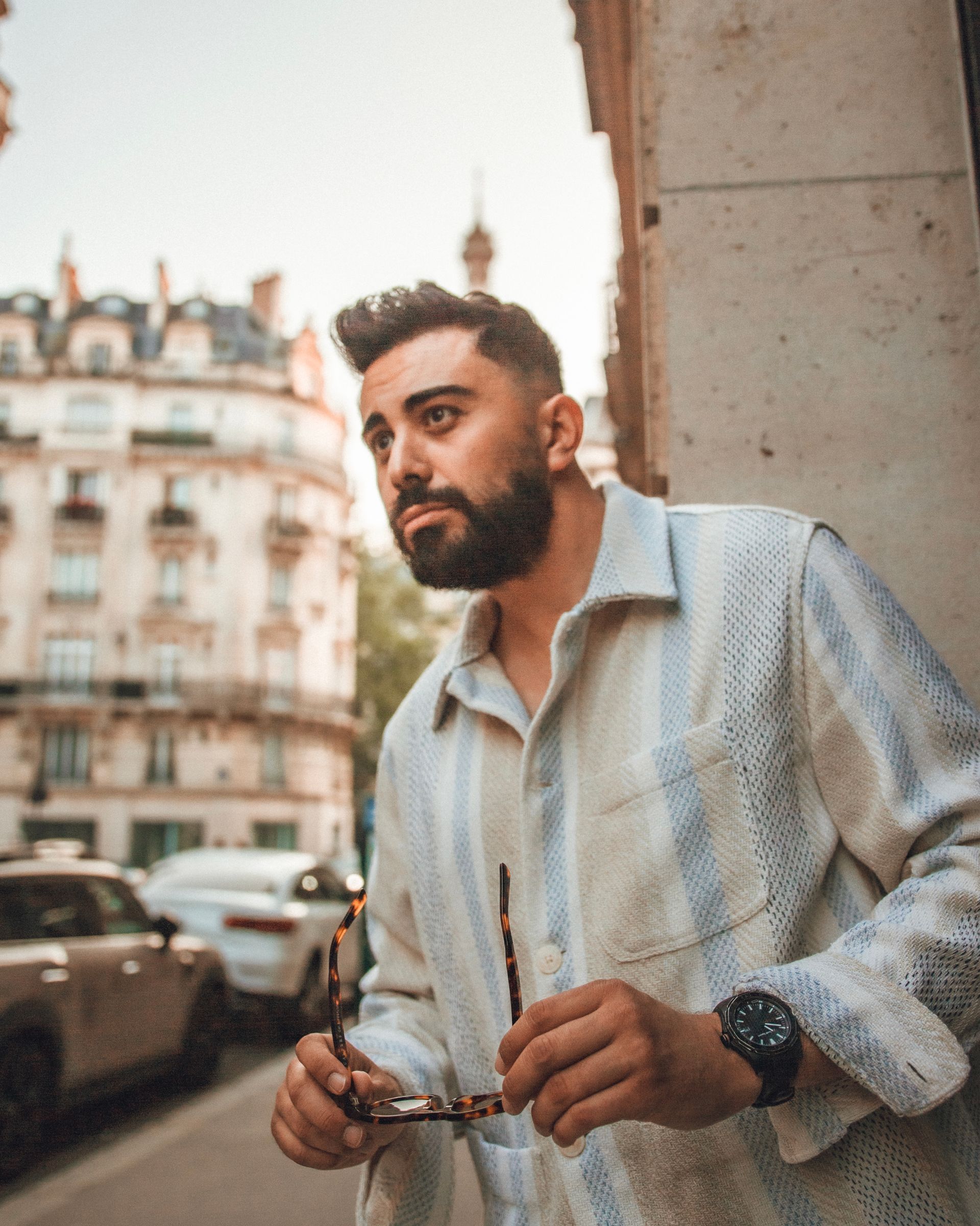 Un homme barbu en chemise bleu clair, tenant des lunettes, se tient dans une rue parisienne avec des bâtiments et des .