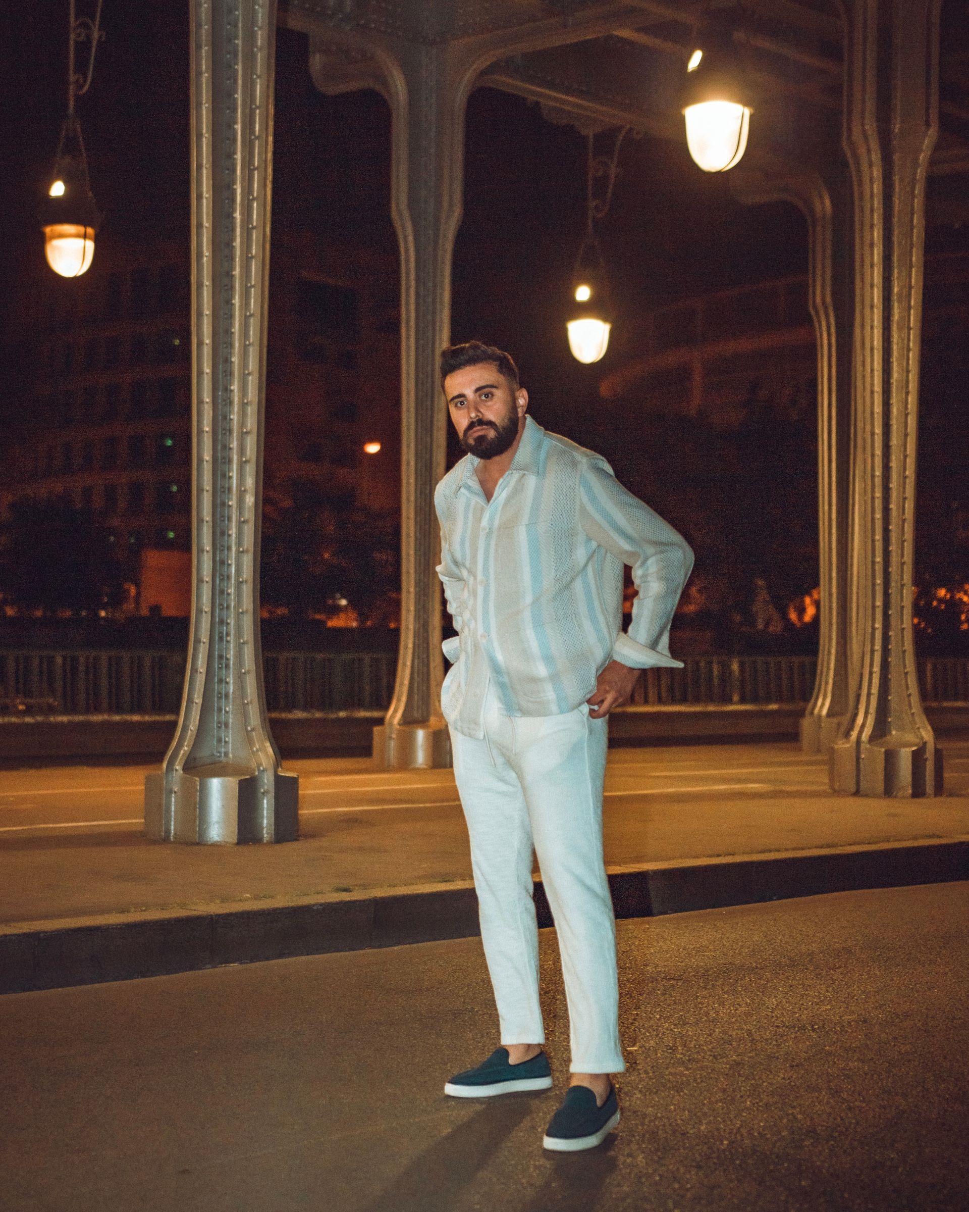 Un homme en tenue blanche se tient dans la rue la nuit, sous un pont avec des lumières suspendues.