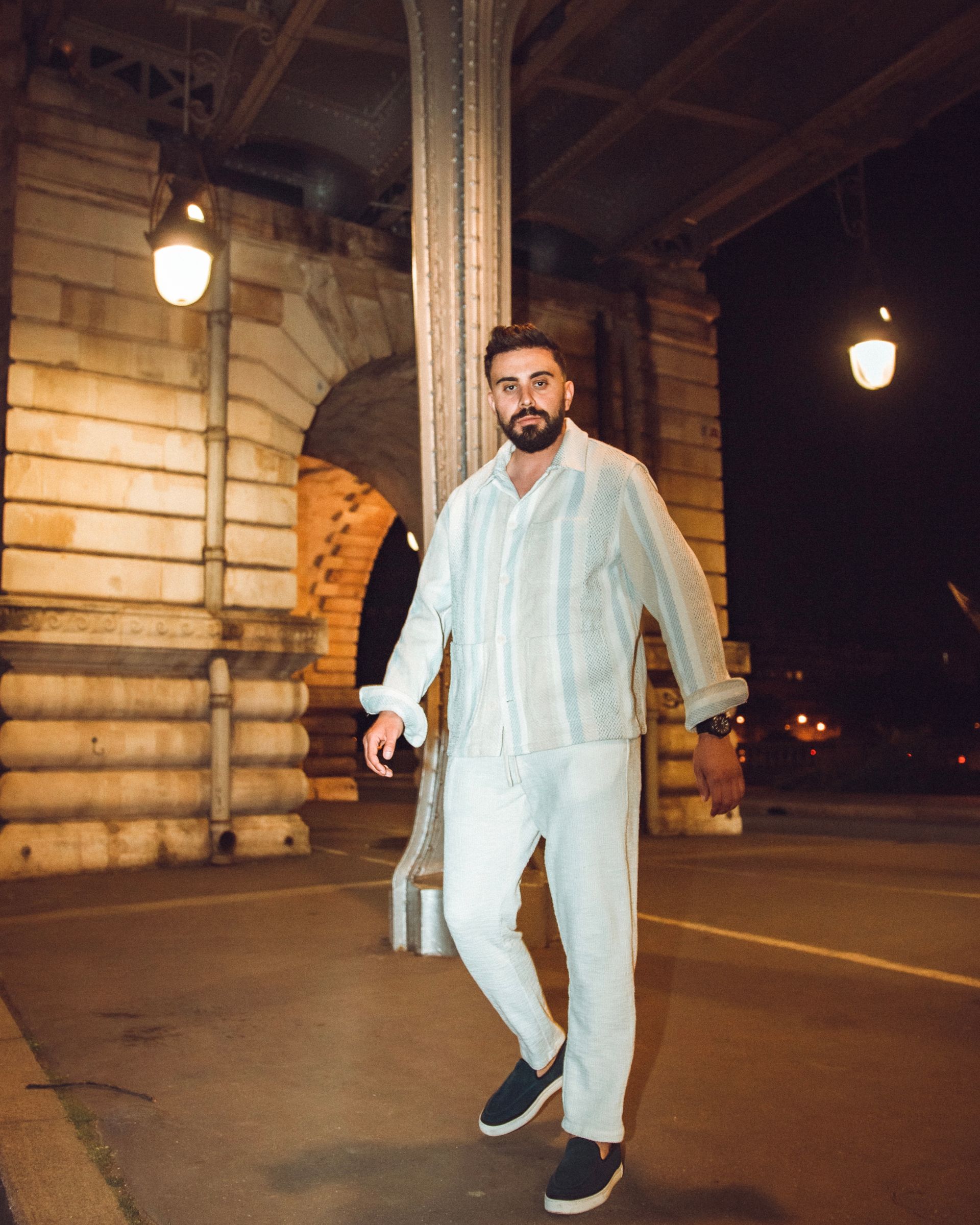 Un homme en tenue de couleur claire marche sur un pont la nuit ; Paris.