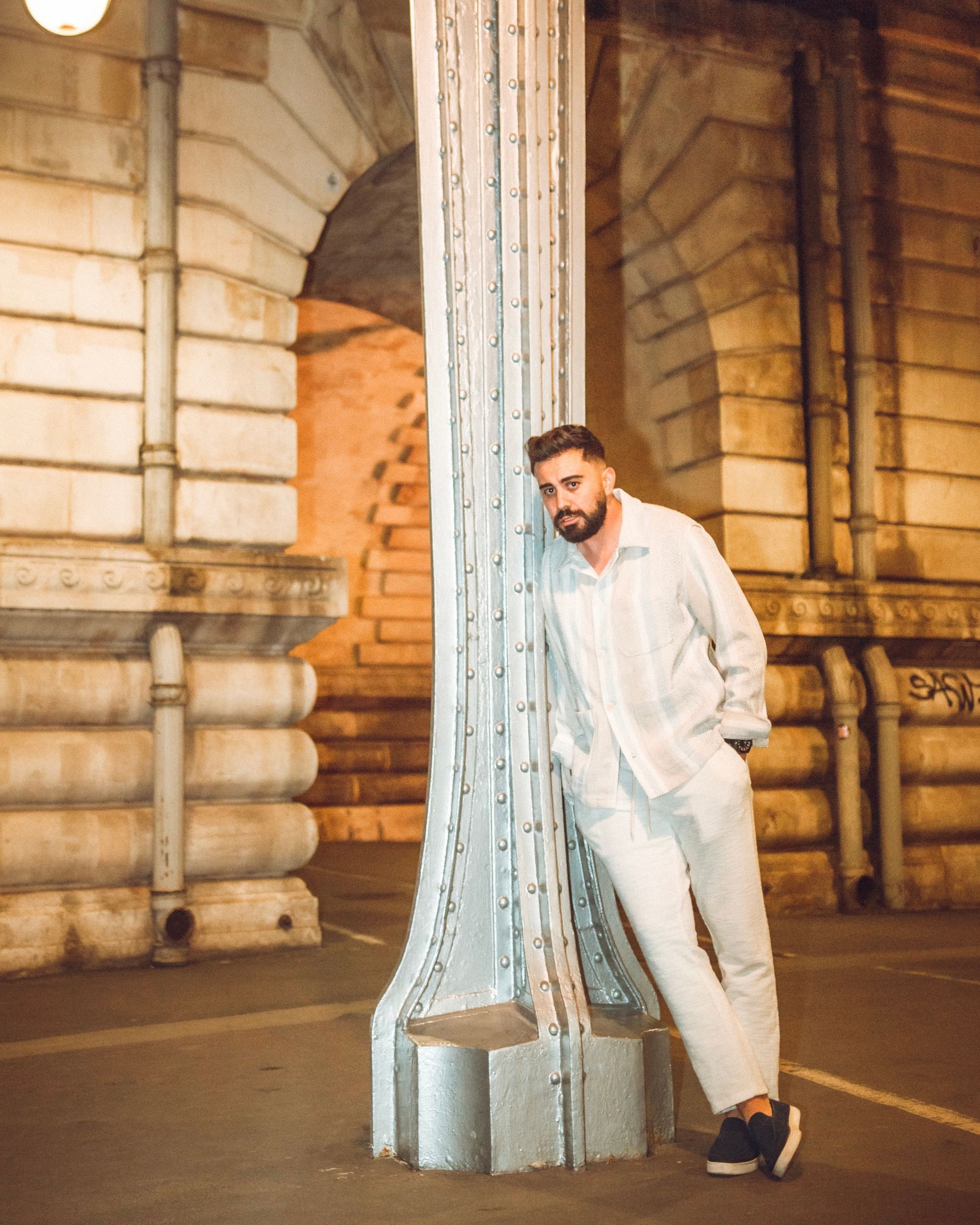 Un homme s'appuie contre une colonne texturée la nuit, vêtu d'une tenue en lin blanc. L'architecture est en arrière-plan.