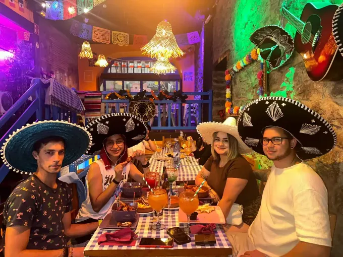 Personas con sombreros en una mesa decorada de un restaurante mexicano. Se ven bebidas, comida y una decoración colorida.