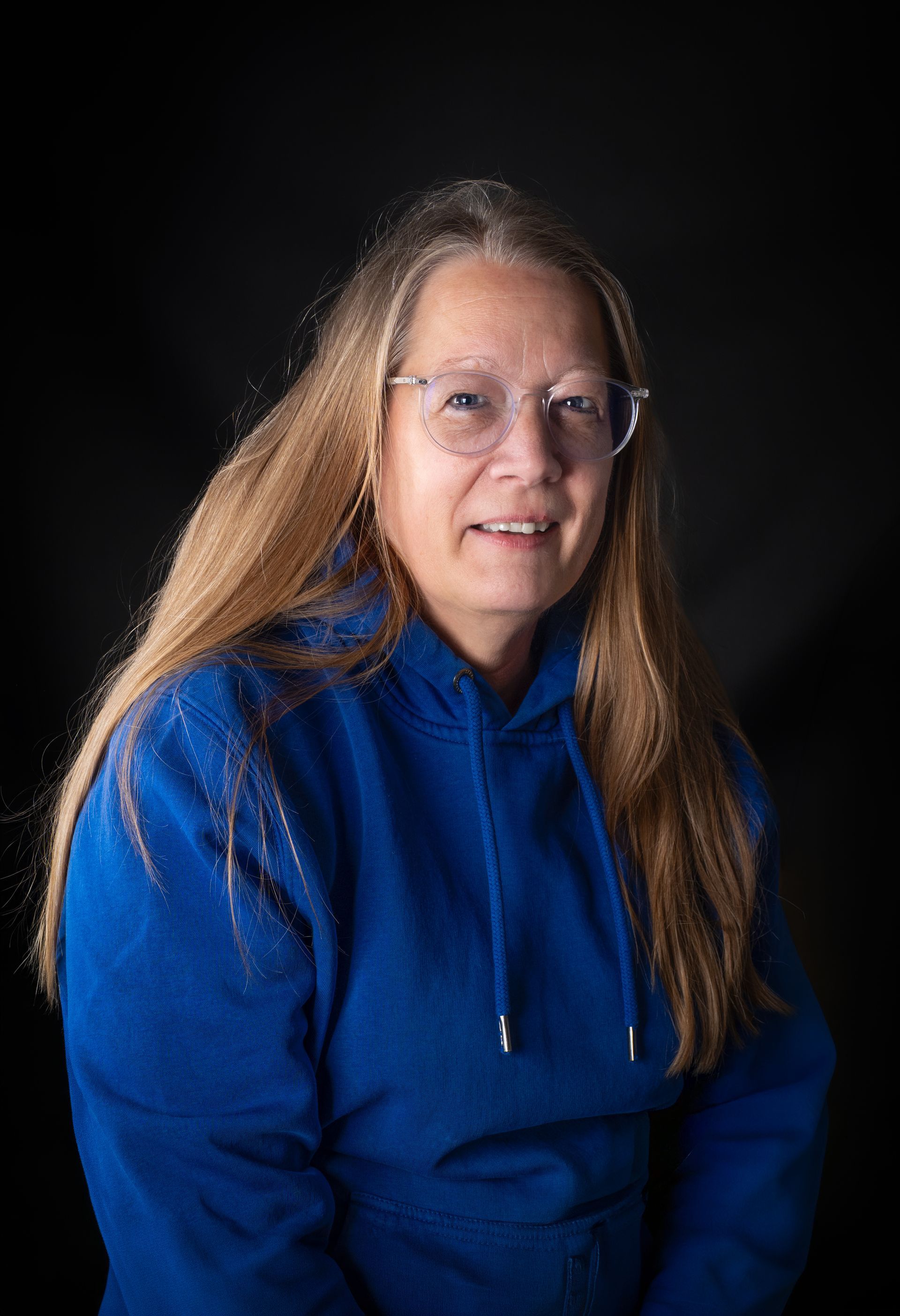 Person in blauem Kapuzenpulli und Brille vor schwarzem Hintergrund, Blick nach vorn gerichtet mit einem leichten Lächeln.