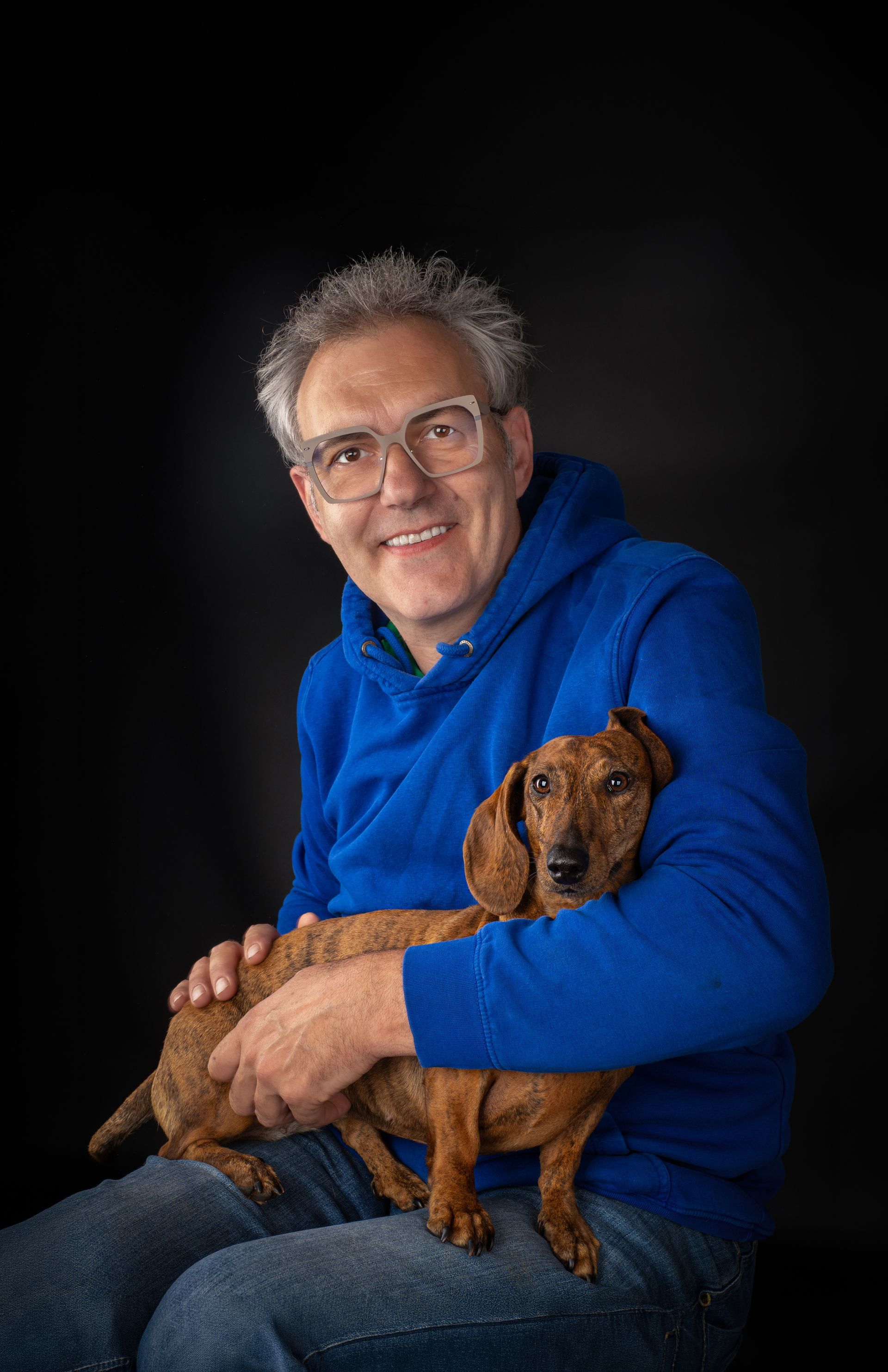 Mann in blauem Pullover hält einen braunen Hund vor dunklem Studiohintergrund