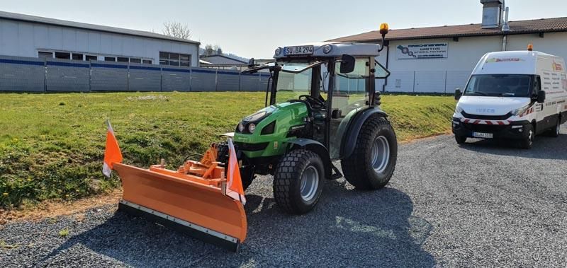Ein grüner Traktor mit einem orangefarbenen Pflug steht neben einem weißen Lieferwagen.