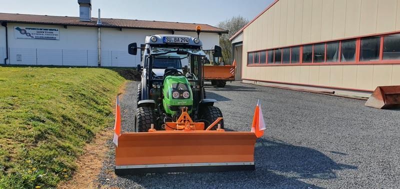 Ein grüner Traktor mit einem orangefarbenen Pflug steht vor einem Gebäude.