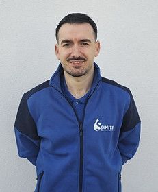 Ein Mann mit einer blauen Jacke und einem blauen Hemd steht vor einer weißen Wand. Sanitek GmbH