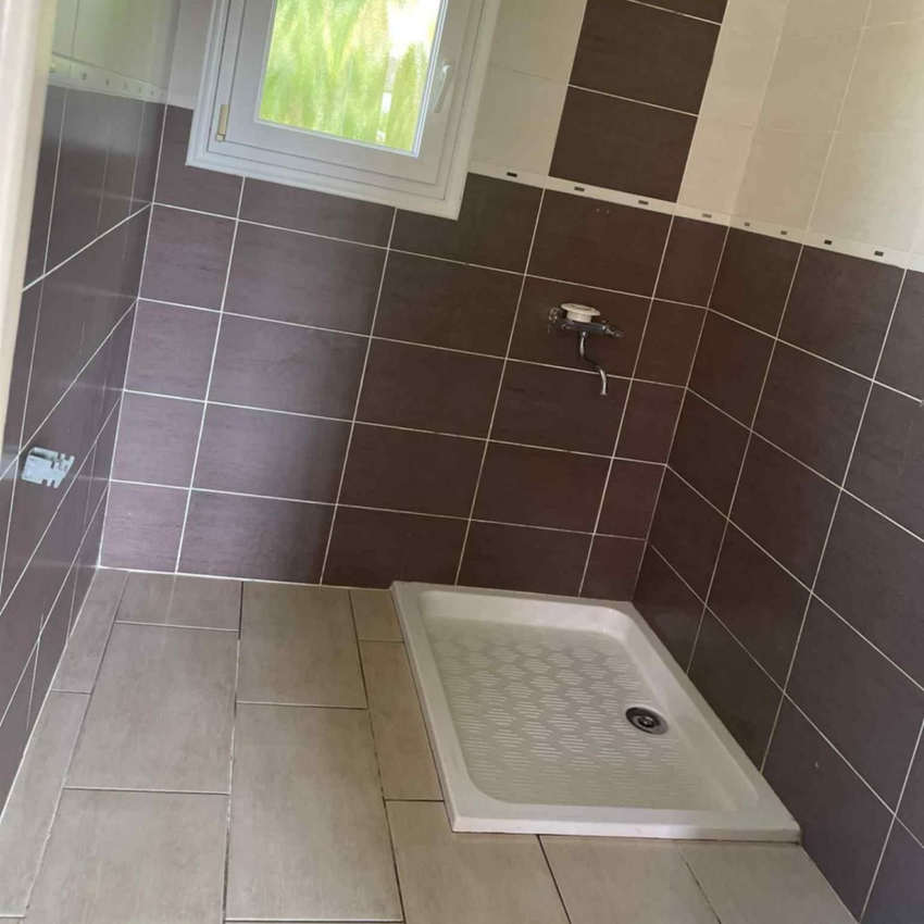 Petite salle de bains carrelée avec receveur de douche, fenêtre et carrelage mural marron et beige.