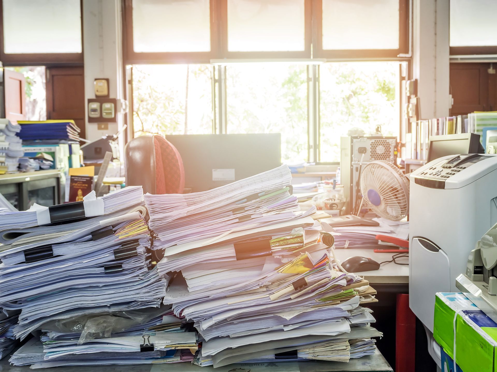 Bureau encombré de piles de papiers et de documents entassés sur les bureaux devant des fenêtres lumineuses.