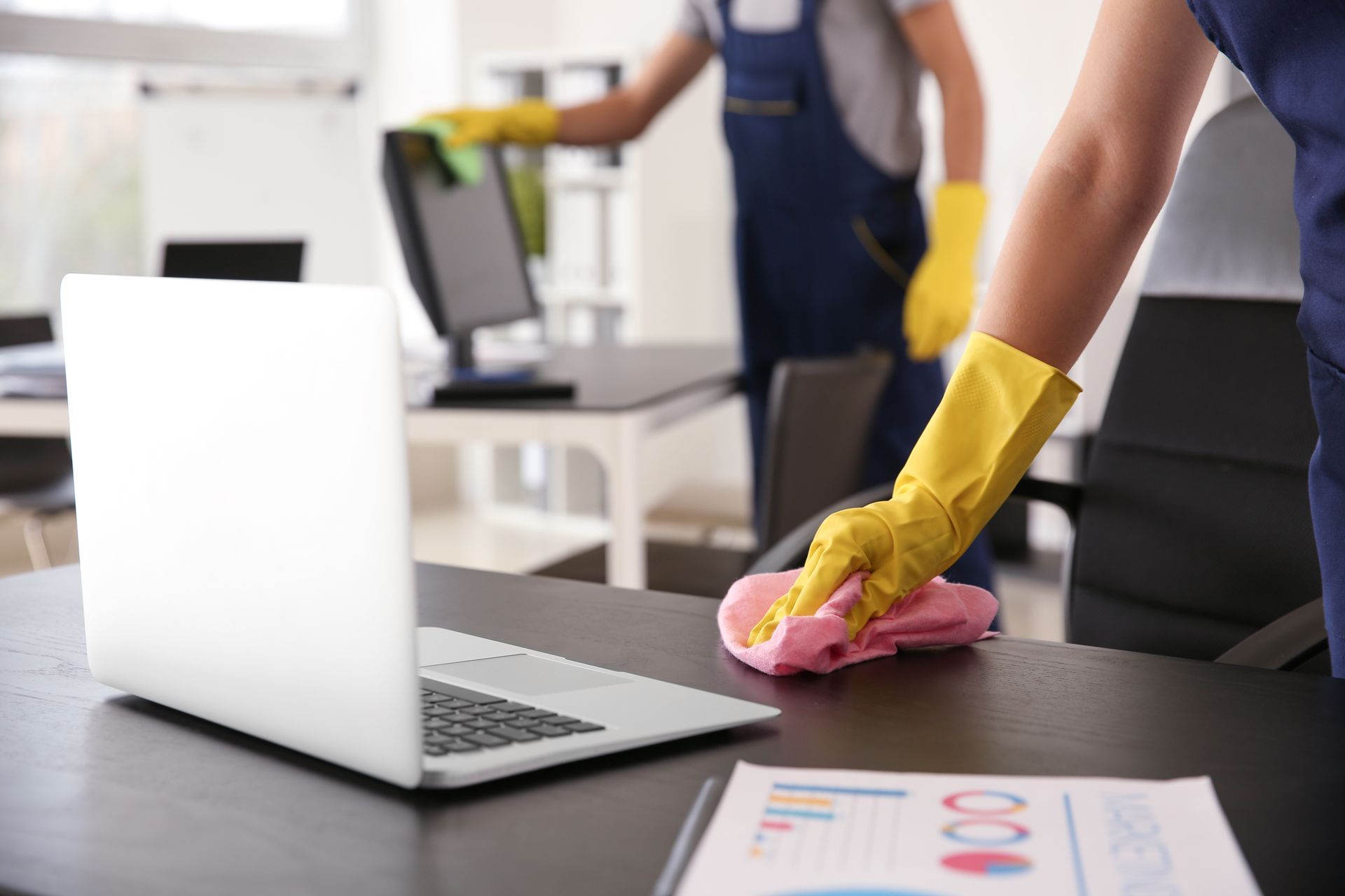 Une personne portant des gants jaunes nettoie un bureau à côté d'un ordinateur portable ouvert dans un bureau.