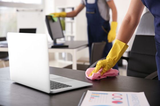Une personne portant des gants jaunes nettoie un bureau à côté d'un ordinateur portable ouvert dans un bureau.