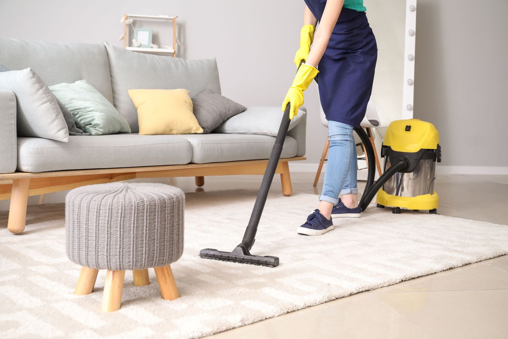 Une personne passe l'aspirateur sur un tapis blanc dans un salon lumineux avec un aspirateur traîneau jaune.