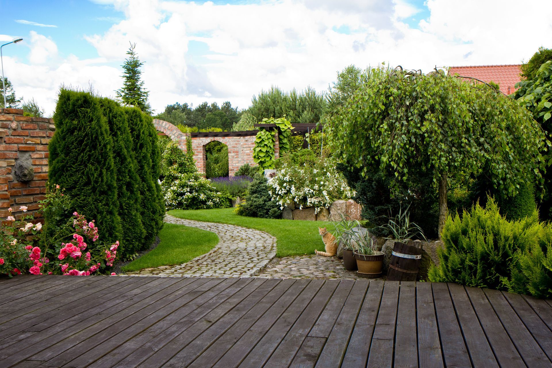 Jardin luxuriant avec terrasse en bois, chemin de pierre sinueux, fleurs, arbustes et arche en briques sous un ciel lumineux.