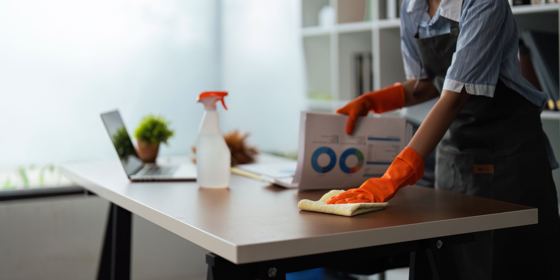Une personne portant des gants orange nettoie un bureau avec un spray et un chiffon.