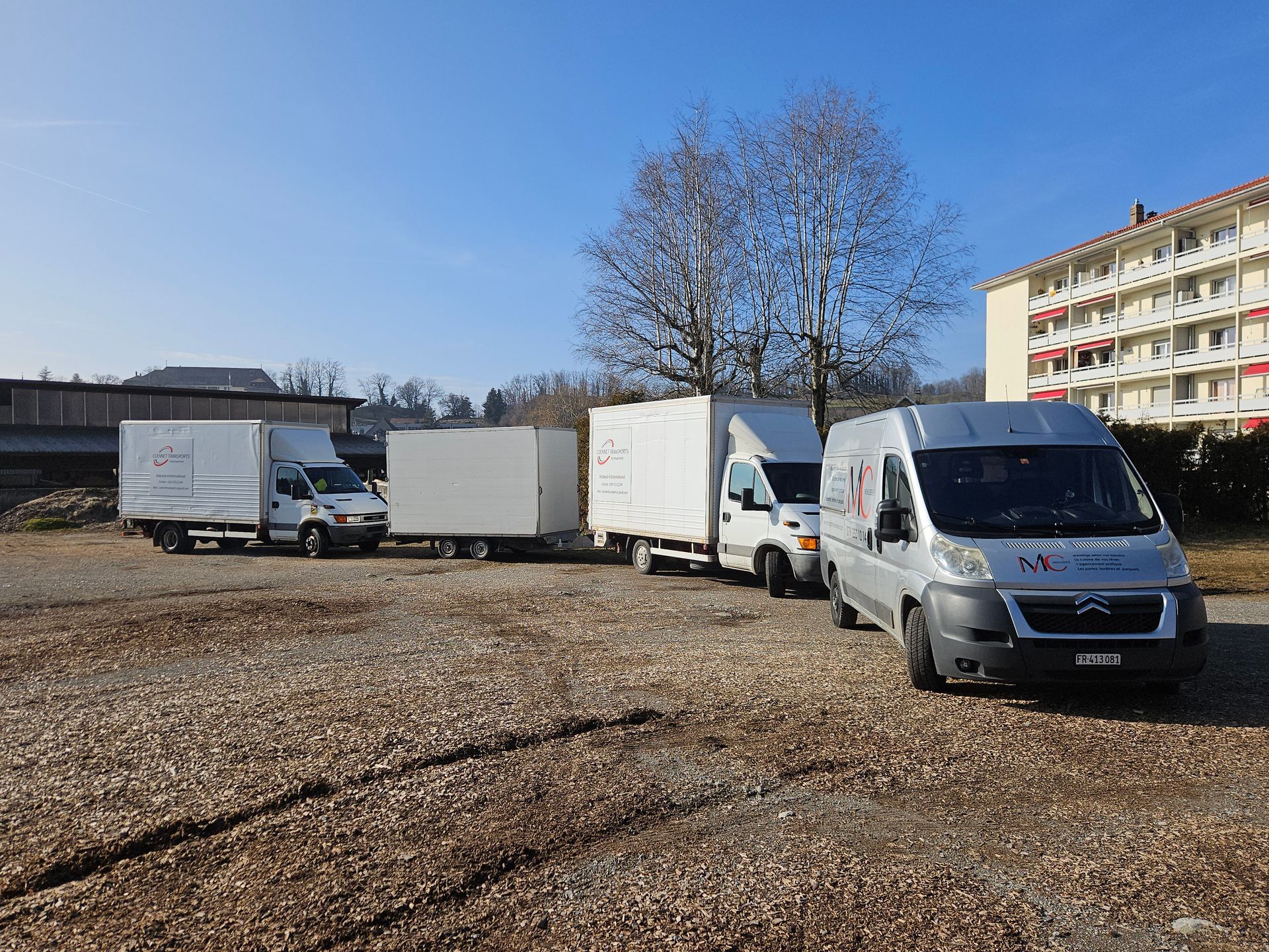 Quatre camionnettes de livraison garées sur un terrain en gravier par une journée ensoleillée.