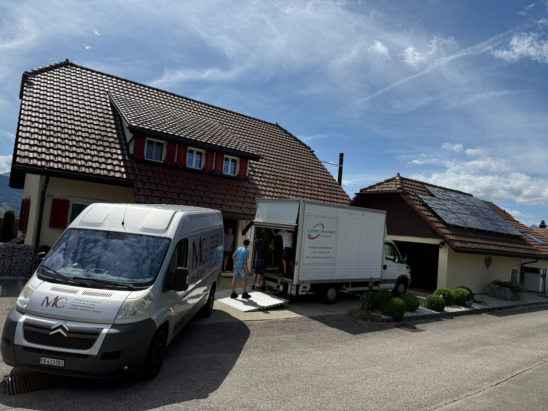 Deux camions de déménagement garés près d'une maison ; des gens chargent des meubles. Journée ensoleillée.
