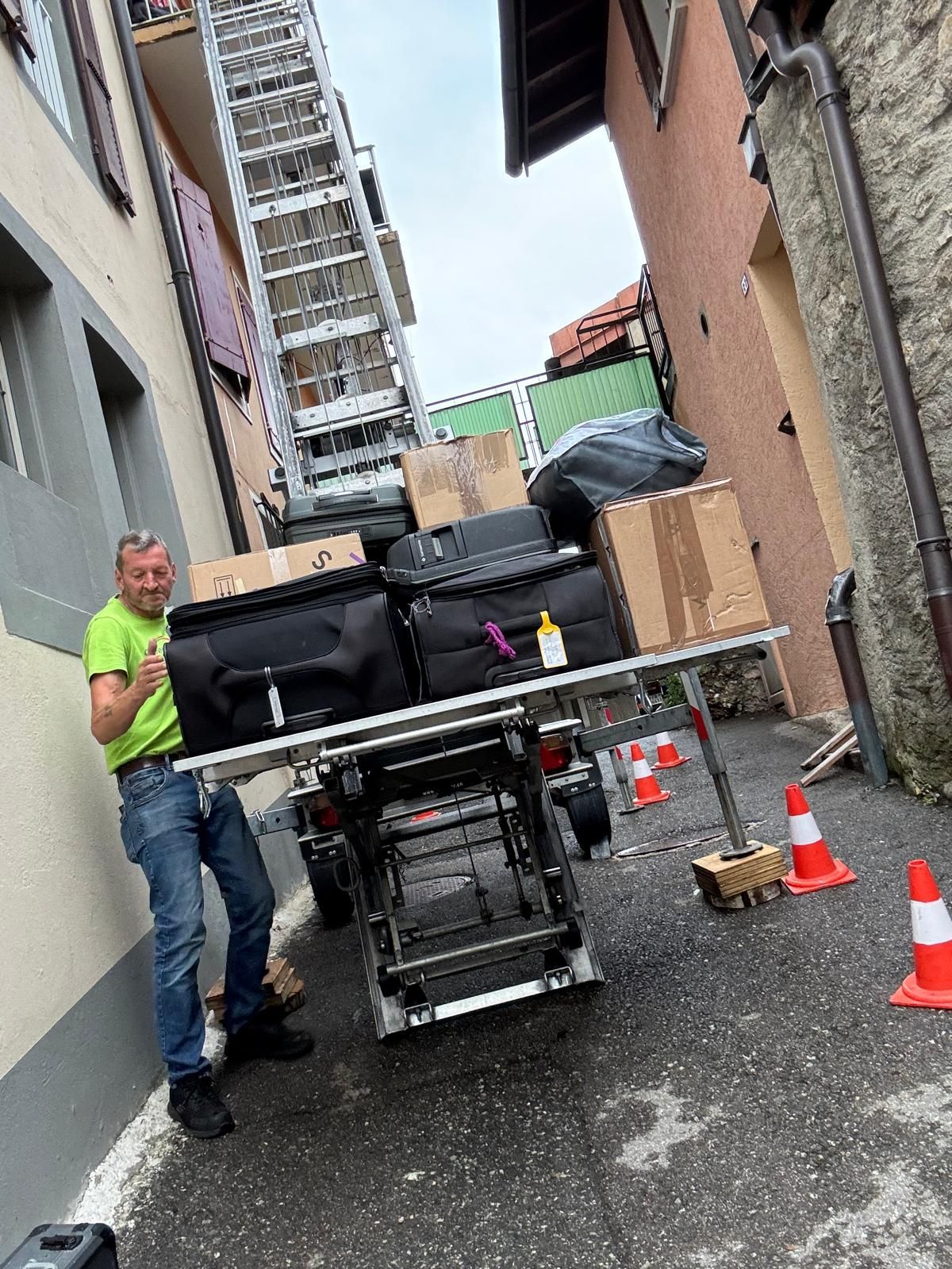 Un homme déplace un ascenseur avec des cartons dans une ruelle. Il lève le pouce. Échelle, cônes rouges, bâtiments.